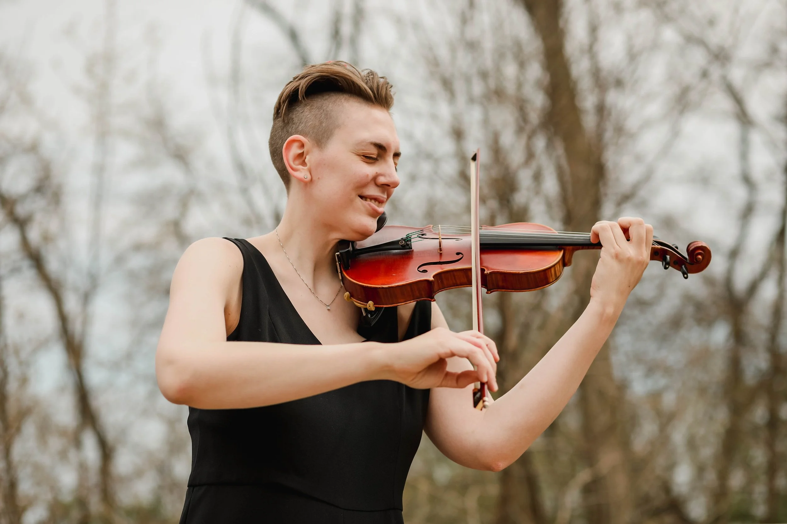 Miss Grube with short hair wearing a black dress playing a violin outdoors among trees.