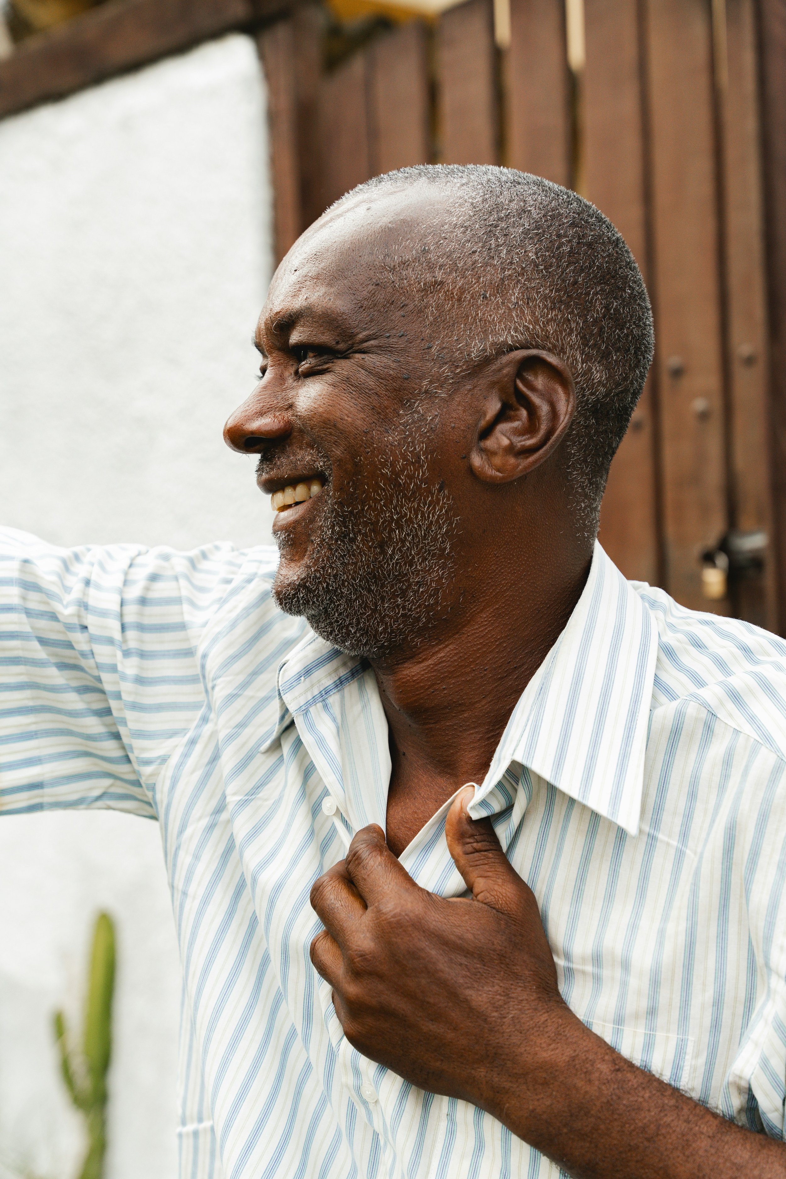 A smiling older man with gray hair and a beard, wearing a striped button-up shirt, adjusting his shirt collar with one hand while outdoors in front of a wooden fence.