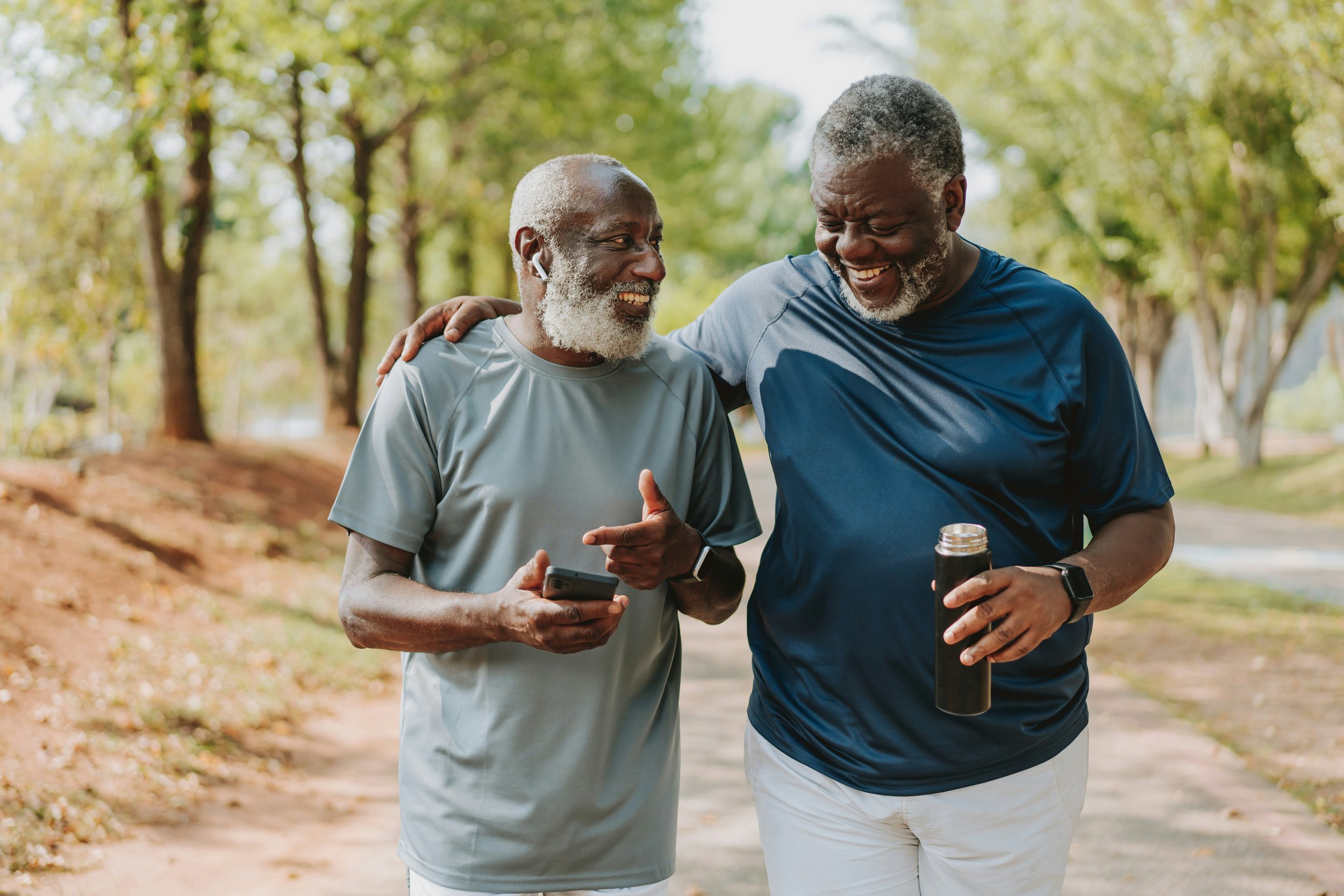 Two elderly men walking in a park, smiling, with one holding a smartphone and the other holding a water bottle, engaging in conversation.