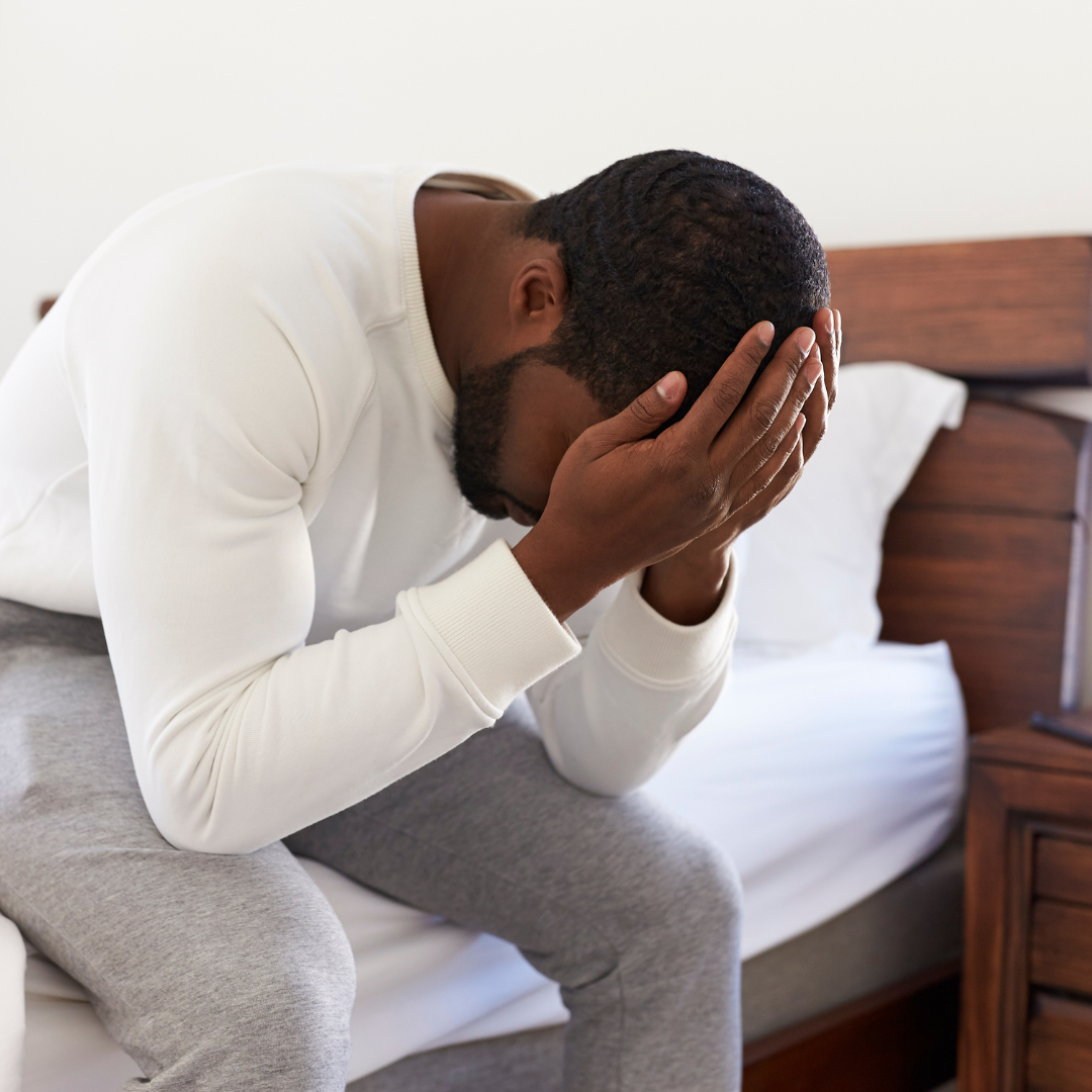 Man sitting on bed, holding his head in his hands, appearing stressed or distressed.