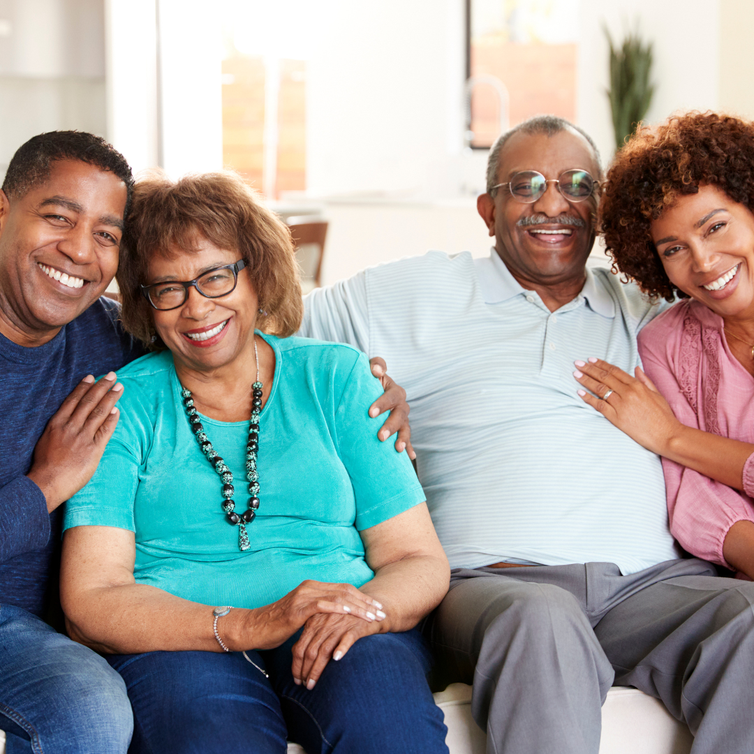 Group of four diverse adults smiling and sitting close together on a couch in a brightly lit living room.