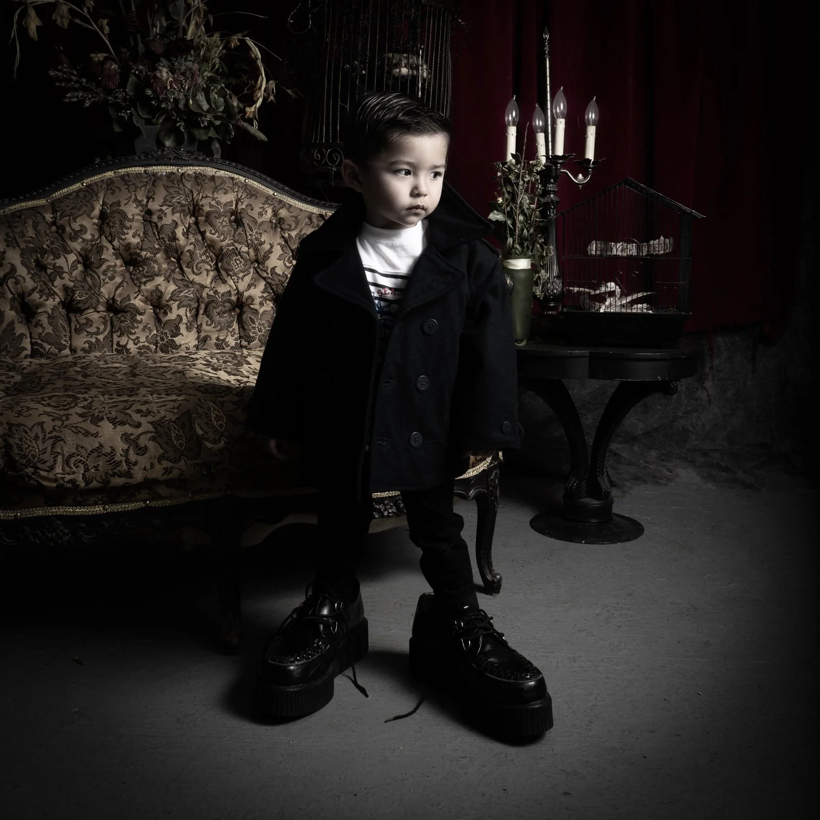 A young boy with slicked-back hair, wearing a black coat, striped shirt, black pants, and oversized black platform shoes, standing in a dimly lit vintage room with ornate furniture, a candelabra, and dark red curtains.