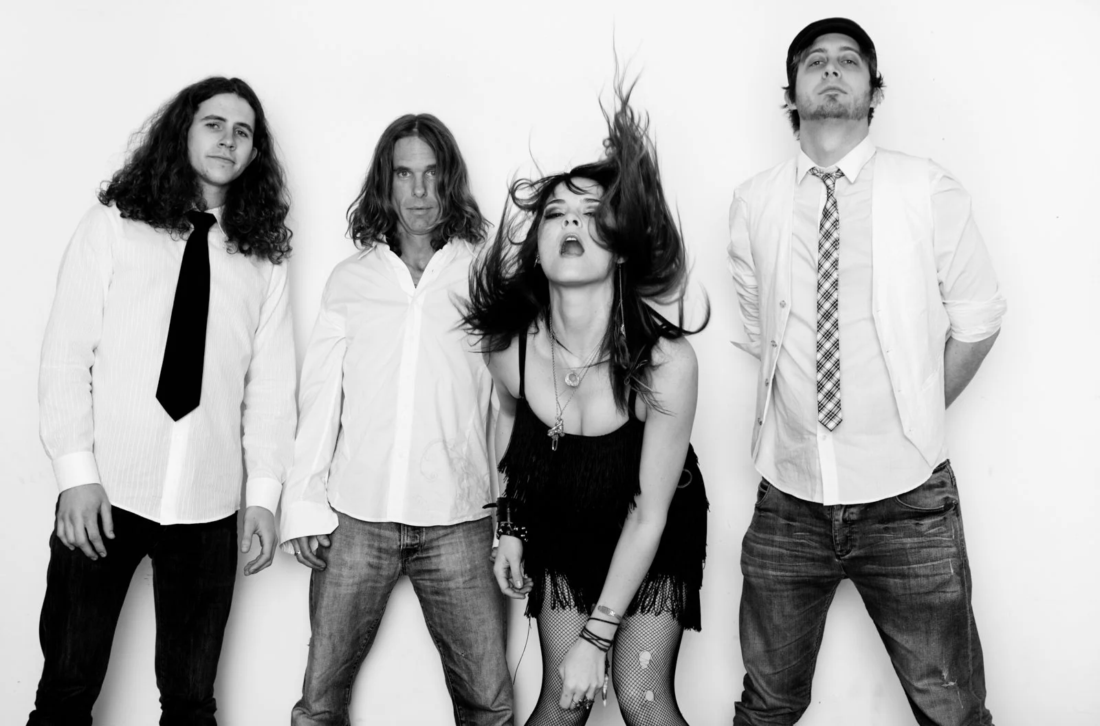 A black and white photo of four people standing against a plain background. The person in the middle has long hair flying and is wearing a sleeveless top and fishnet stockings, with a confident expression. The other three individuals are dressed in c