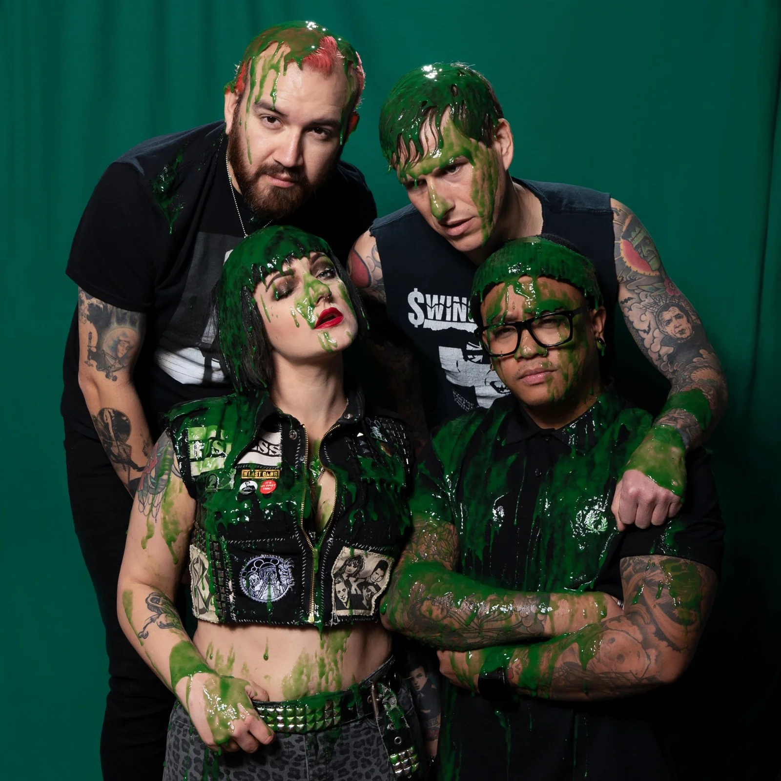 A group of five people covered in green slime, posing in front of a green screen backdrop. Some have tattoos and are wearing punk clothing and accessories.