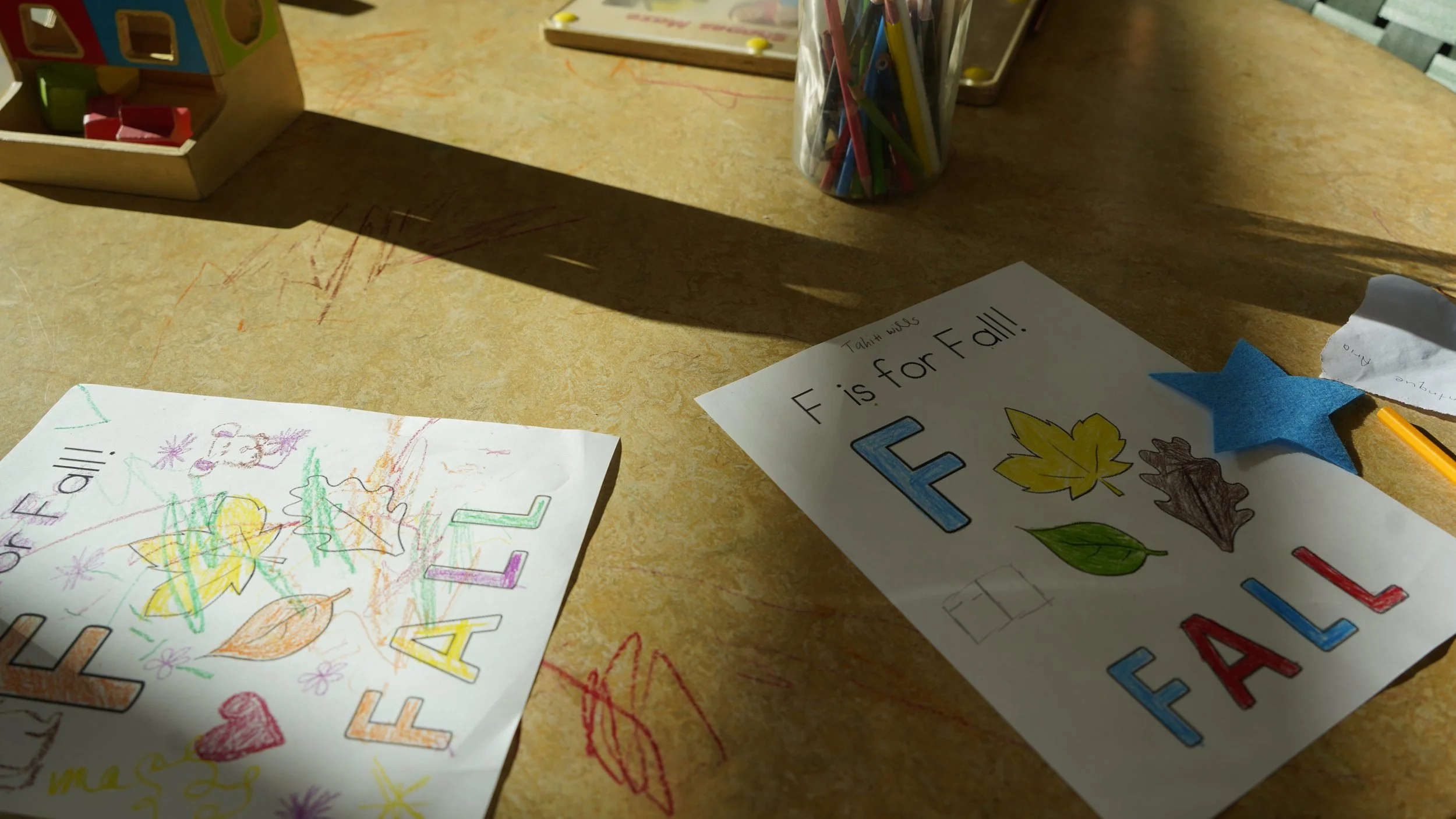 Two children's coloring pages that read "F is for Fall" and colored in different styles with crayon. The pages lay on a light brown table marked with crayon scribbles.