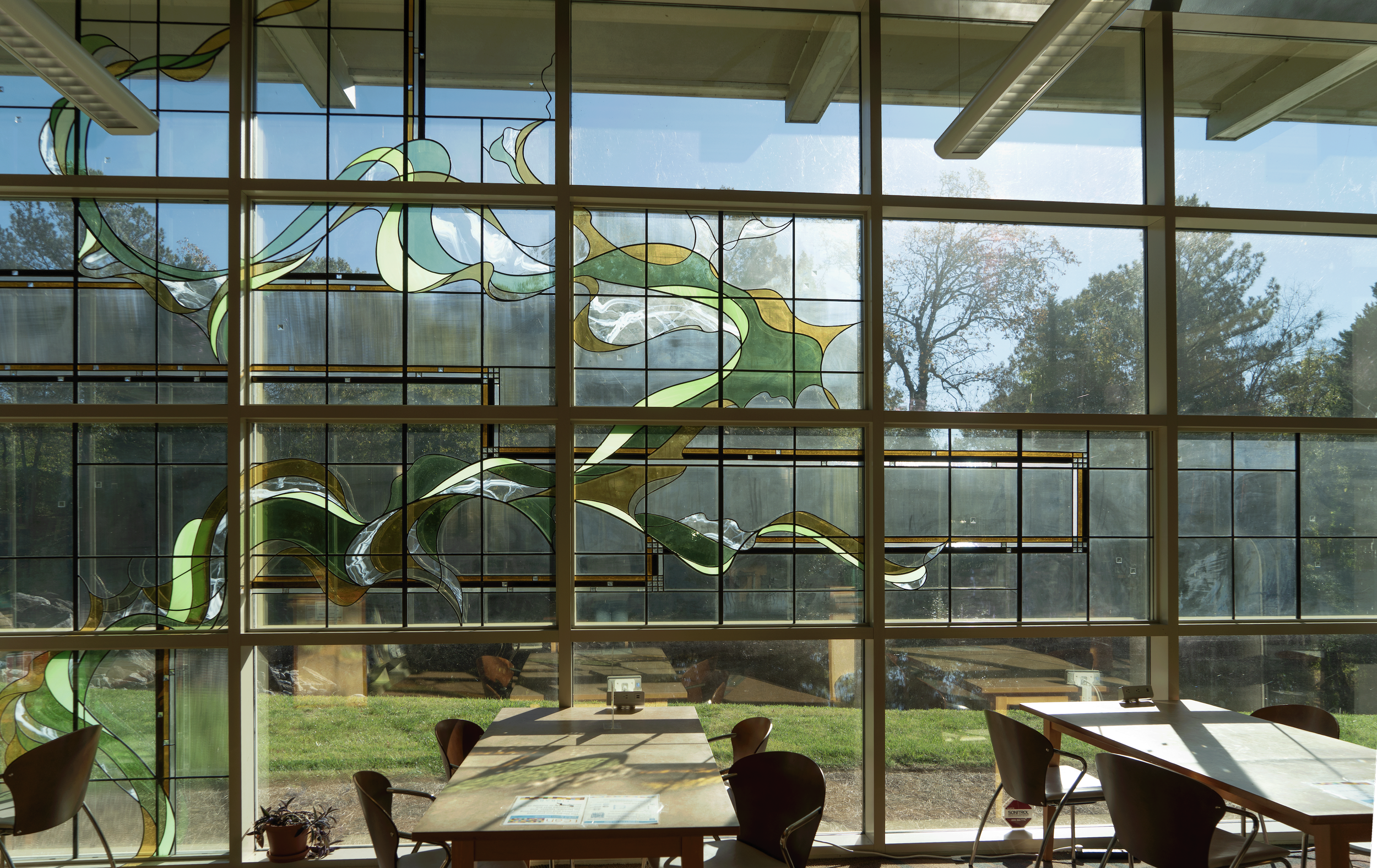 Floor-to-ceiling paned windows with yellow and green art deco stained glass detail. A back patio with a grassy area, a line of trees, and a small creek is visible.