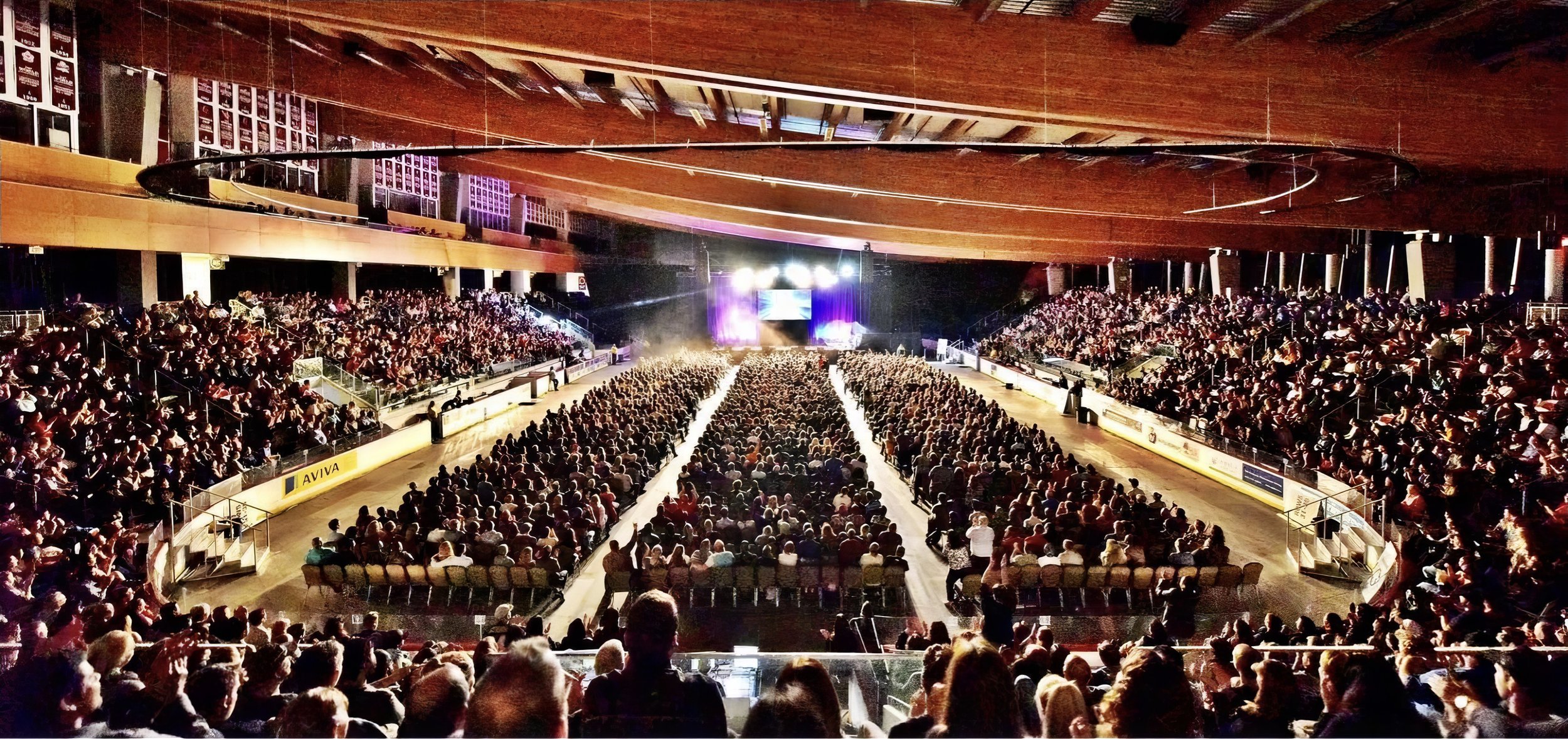 A large indoor arena filled with an audience seated and waiting for a performance on stage, illuminated with colorful lights, with a curved wooden ceiling.