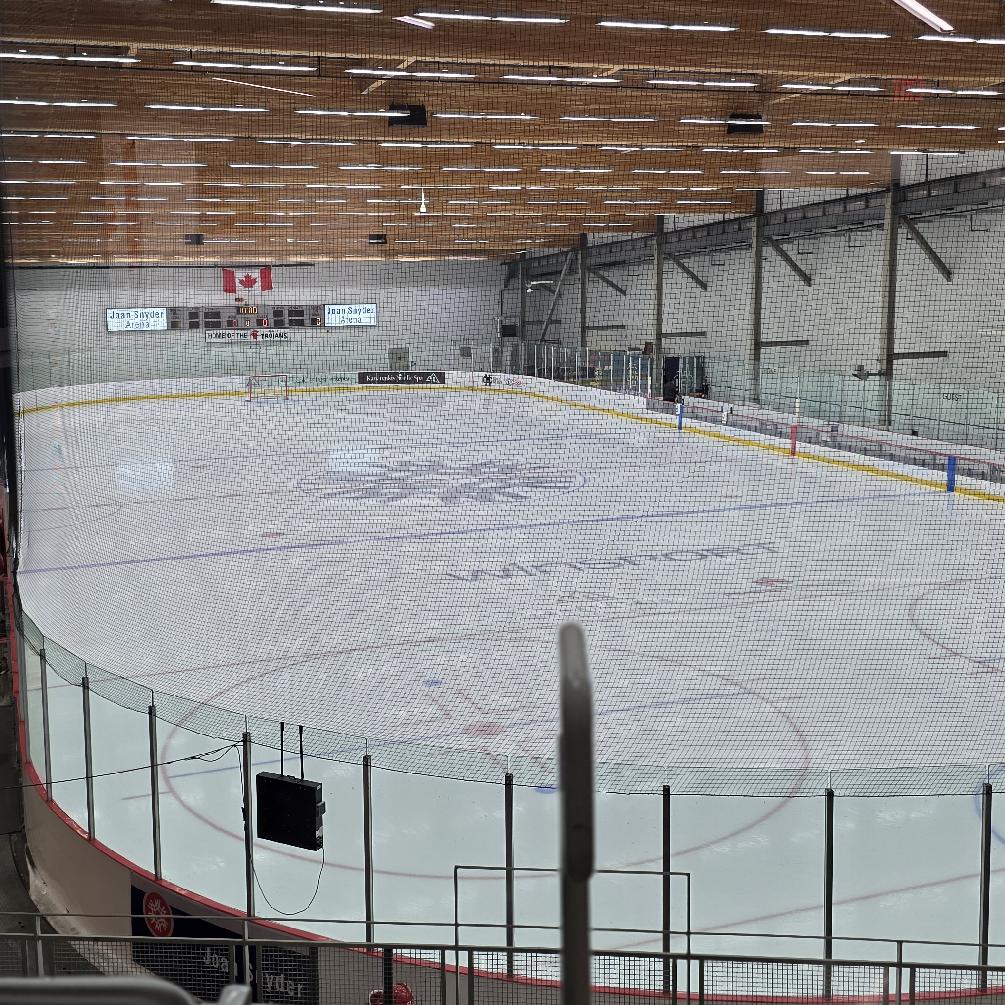 An empty ice hockey rink seen through a protective netting, with a Canadian flag hanging on the wall and a digital scoreboard displaying 10:00.
