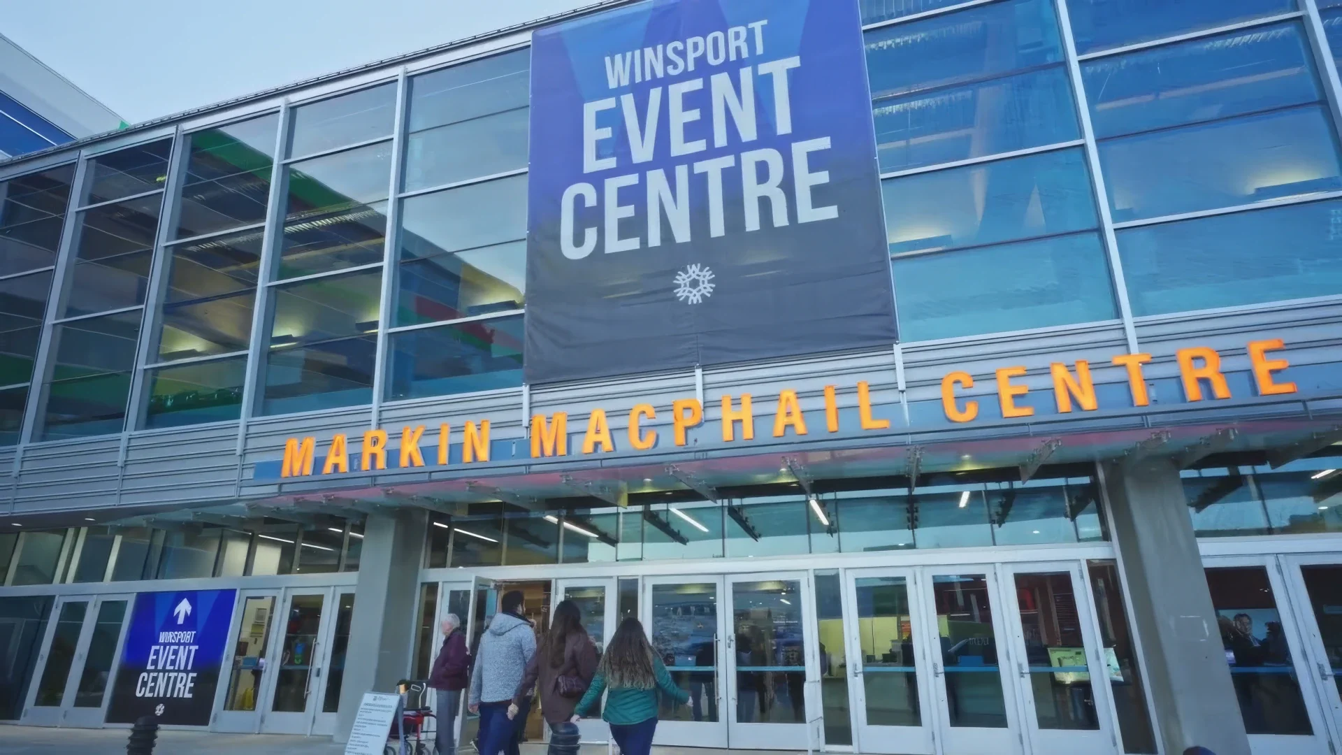 Exterior view of the Winsport Event Centre at Markin MacPhail Centre, with a large banner advertising the event center hanging on the building's glass facade and people entering the building.