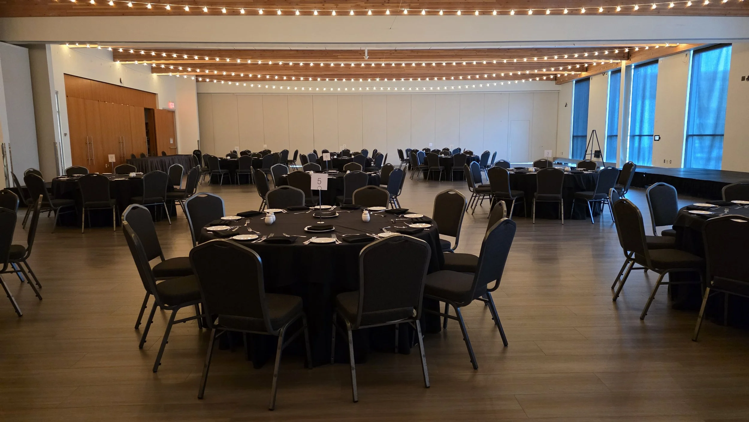 Empty banquet hall set up with round tables covered with black tablecloths, black chairs, place settings, and table numbers, with string lights hanging from the ceiling and large blue curtains on the windows.