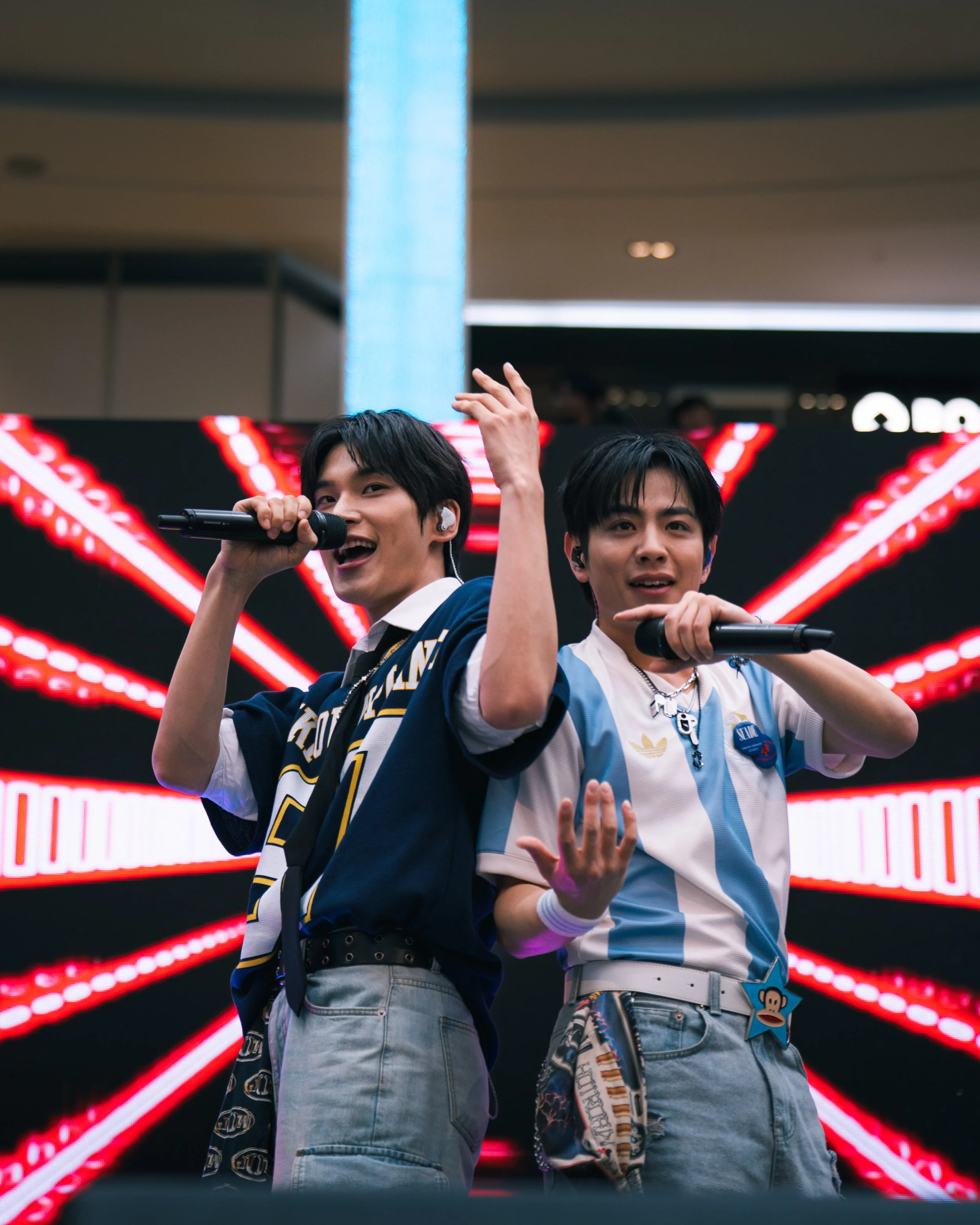 Two young men singing into microphones on stage with a black and red LED display behind them.
