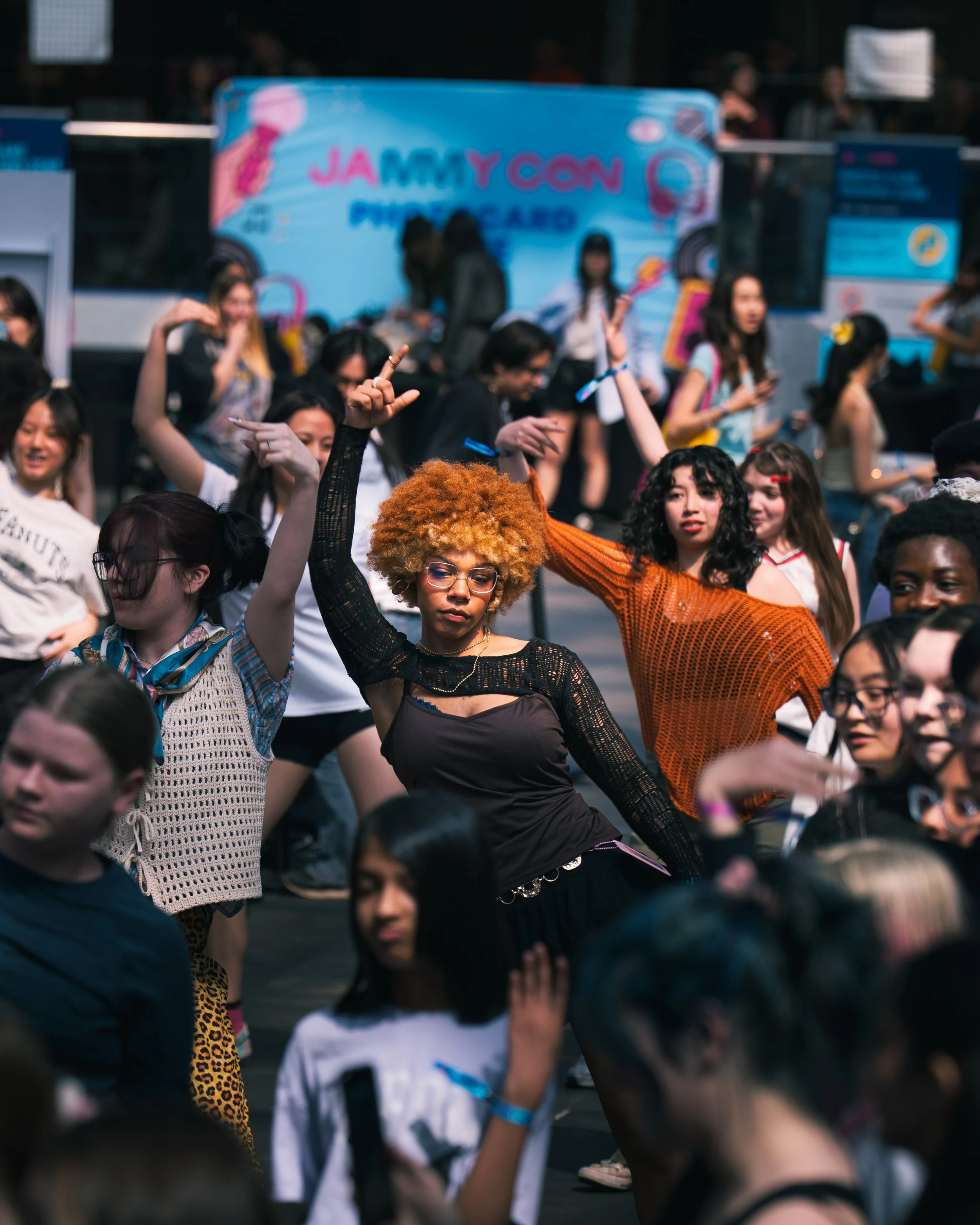 A crowd of young women dancing at an outdoor event with a blue banner in the background.