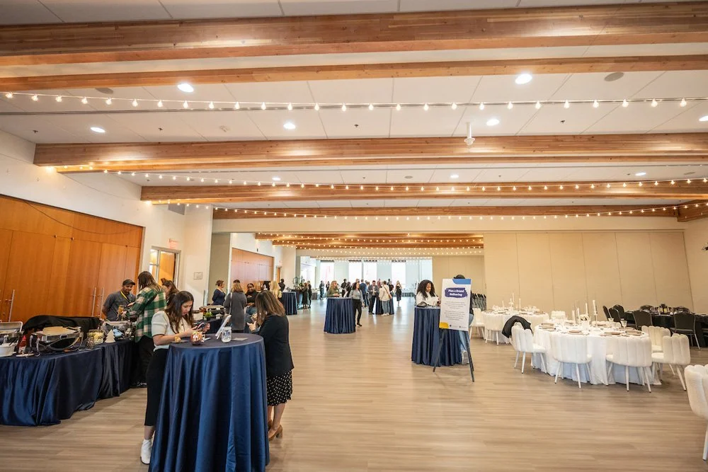 A spacious event hall decorated with string lights, with tables set for a dining event and people mingling around.