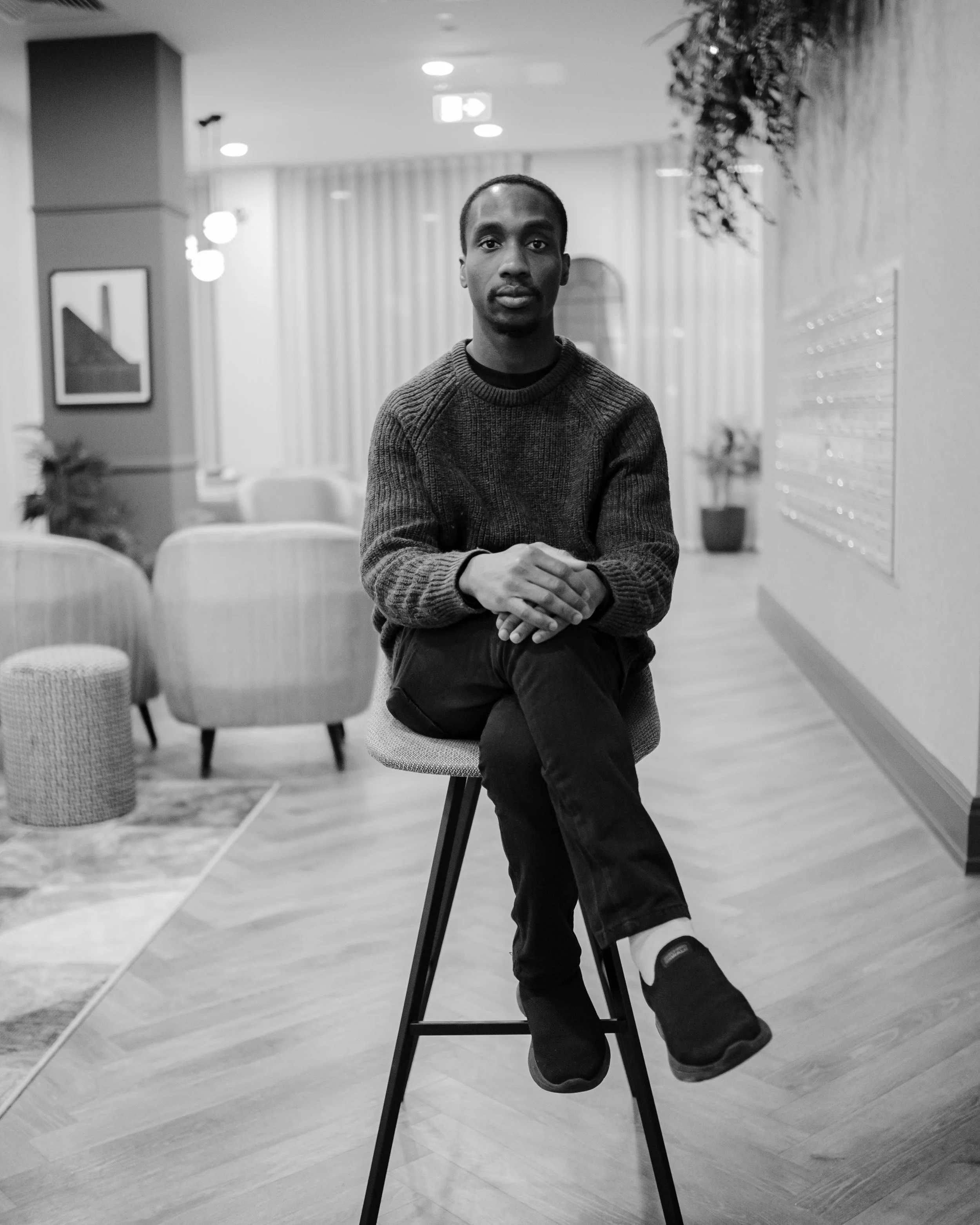 A young man with dark skin and short hair sitting on a high stool in a modern, stylish lounge area, looking directly at the camera. The space features contemporary furniture, artwork, and plants.