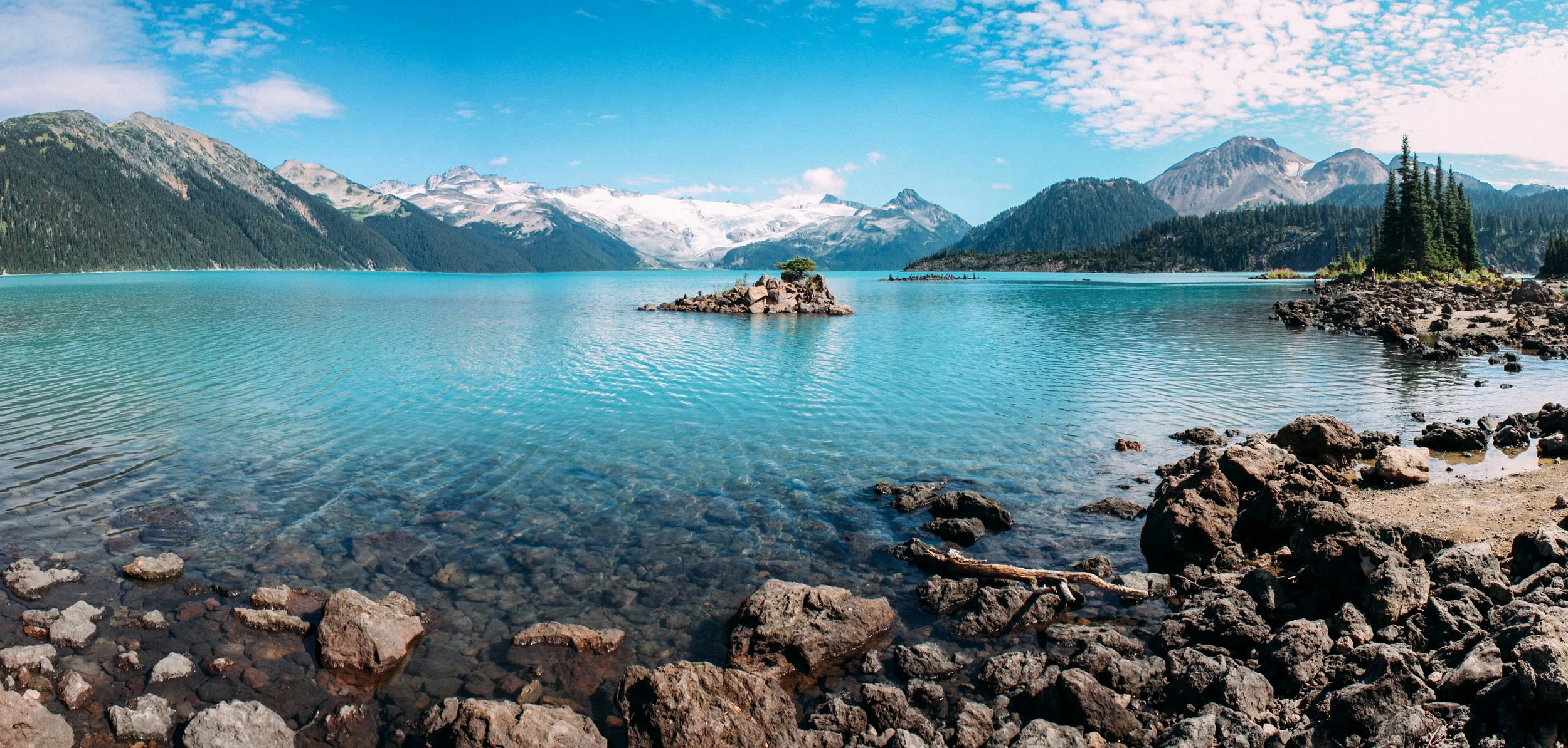A serene mountain lake with clear blue water, surrounded by rocky shores and lush pine trees, with snow-capped mountains and a partly cloudy sky in the background.