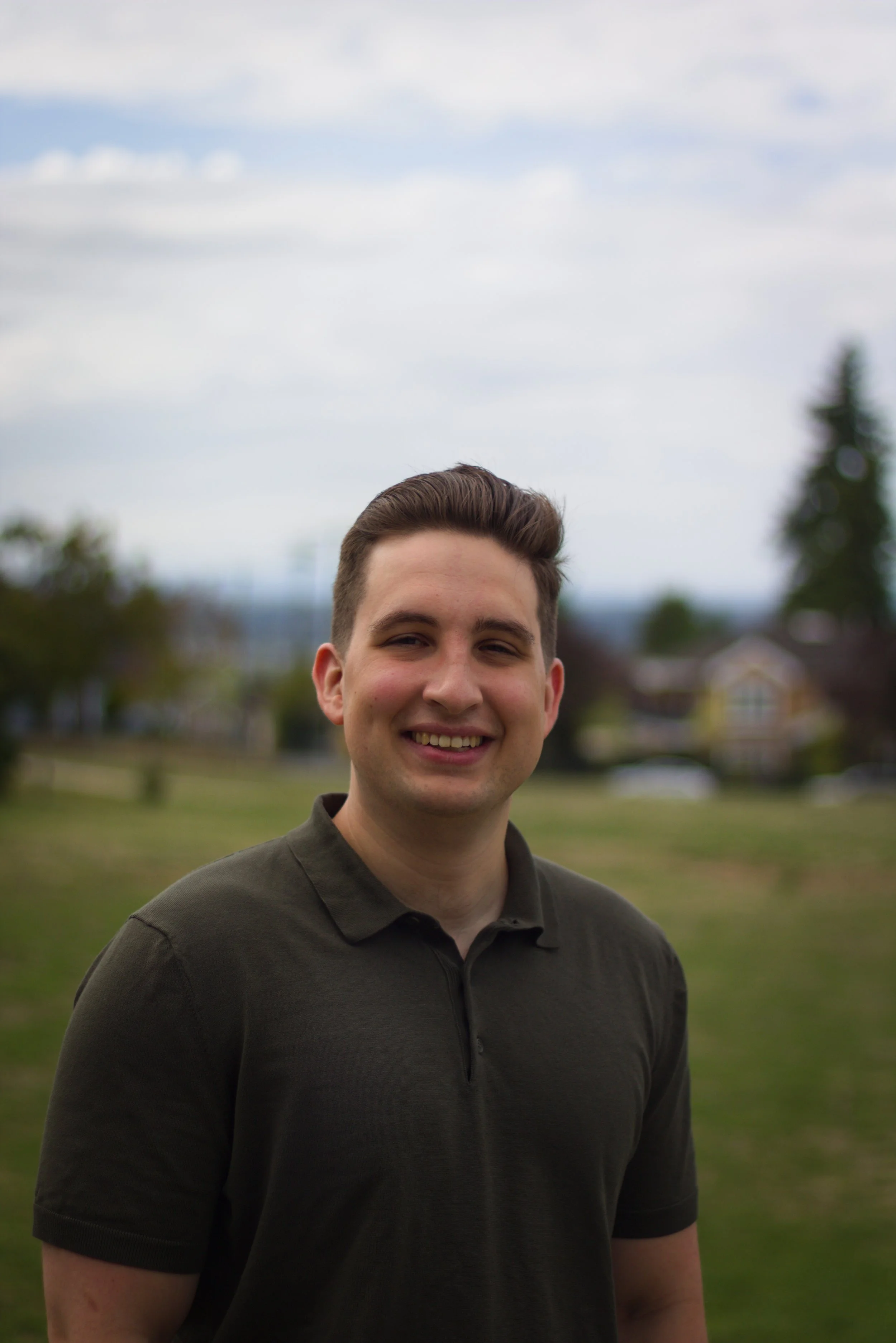 A young man with short brown hair smiling outdoors in a park with green grass and trees, under a cloudy sky.