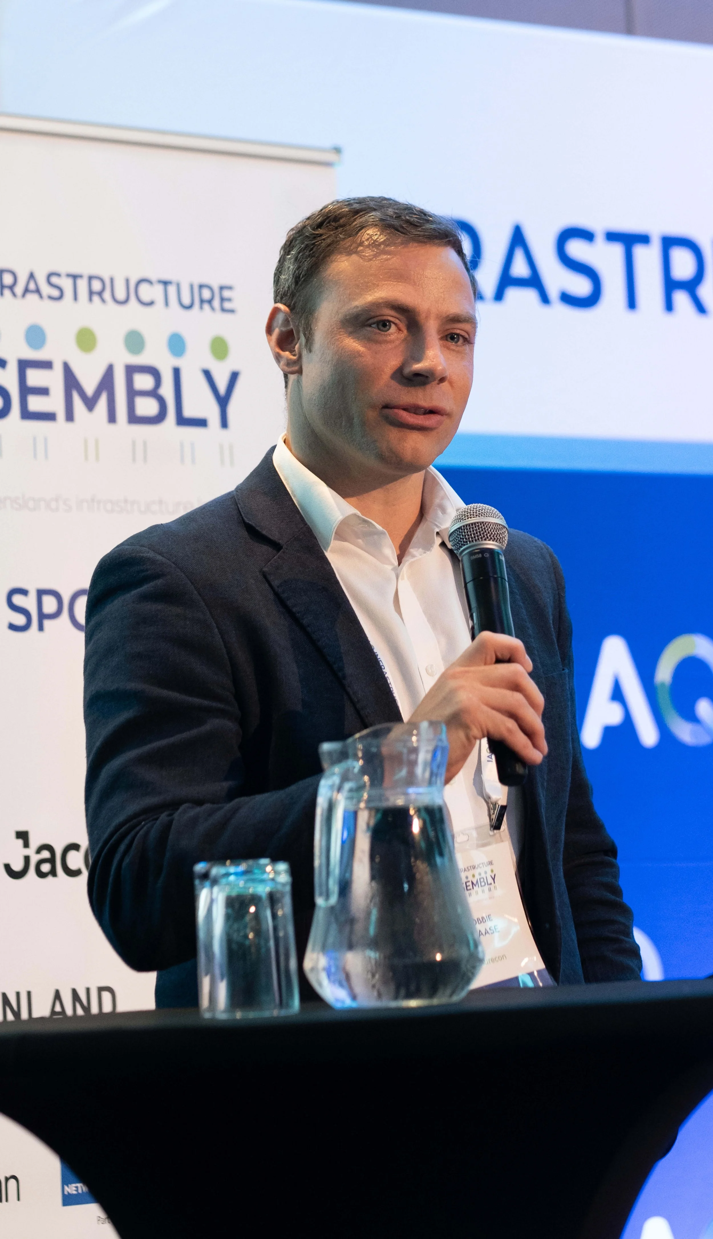 Robbie Claase, in a blazer and white shirt speaking into a microphone at a the IAQ conference, with a blue and white backdrop featuring the words ‘ASSEMBLY’ visible. There is a glass pitcher and glass on the table in front of him.