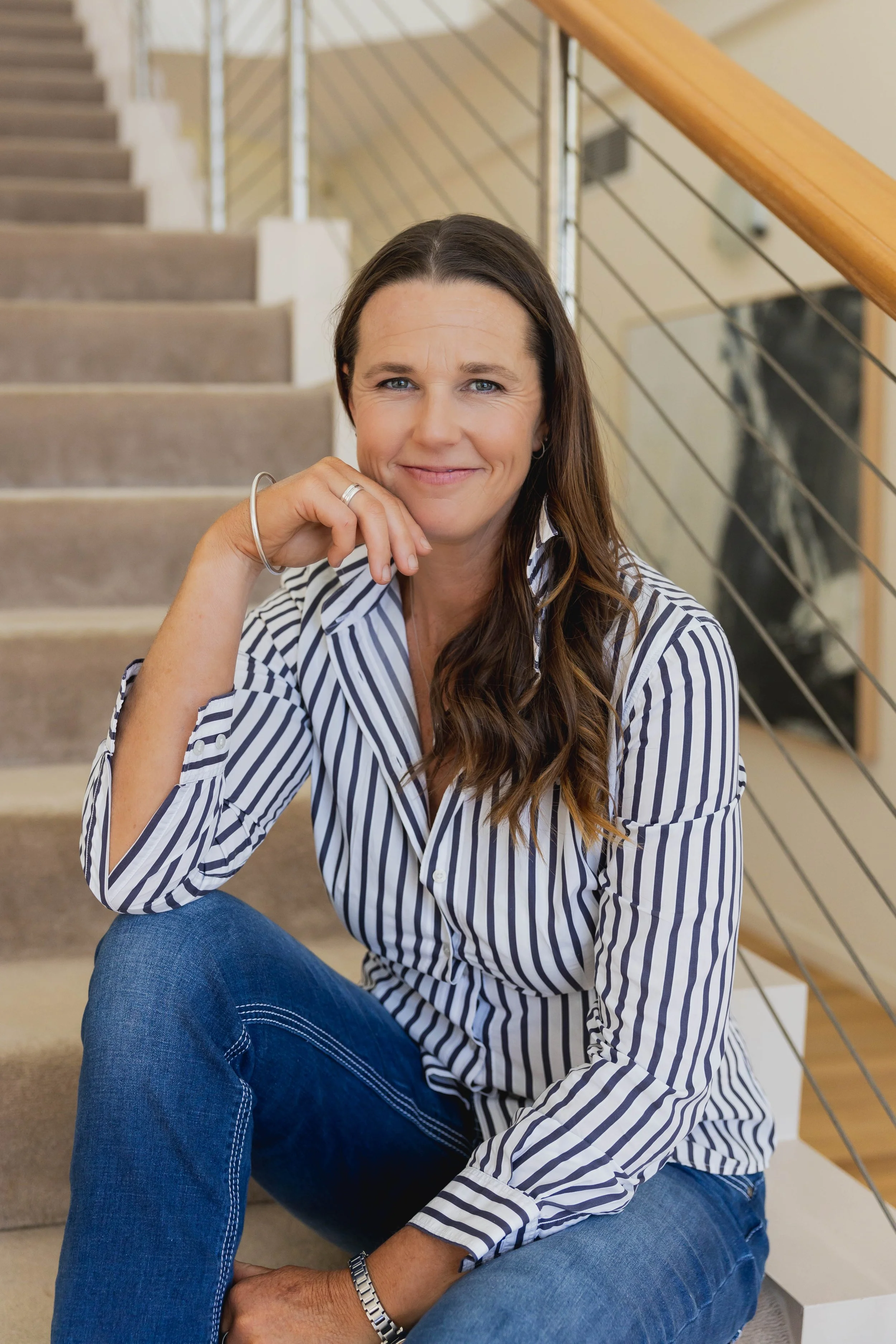 Candid portrait of Lucy Peel, founder of Lucy Peel Human Design, smiling warmly while seated on a staircase, wearing a light-coloured top and silver jewelry.