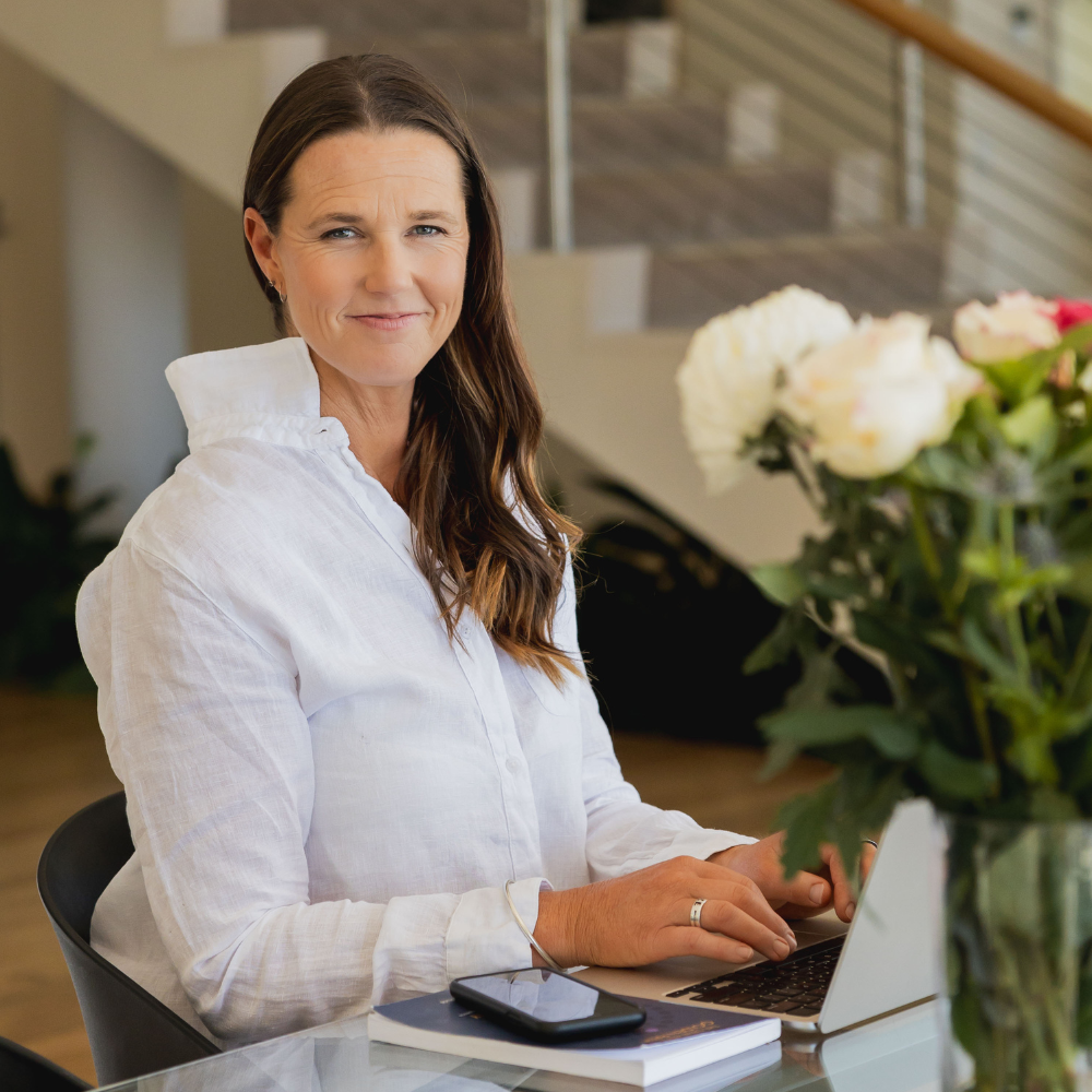 Focus on Lucy Peel, Human Design expert, typing on a laptop computer at a transparent desk with a floral arrangement of white and pink roses.