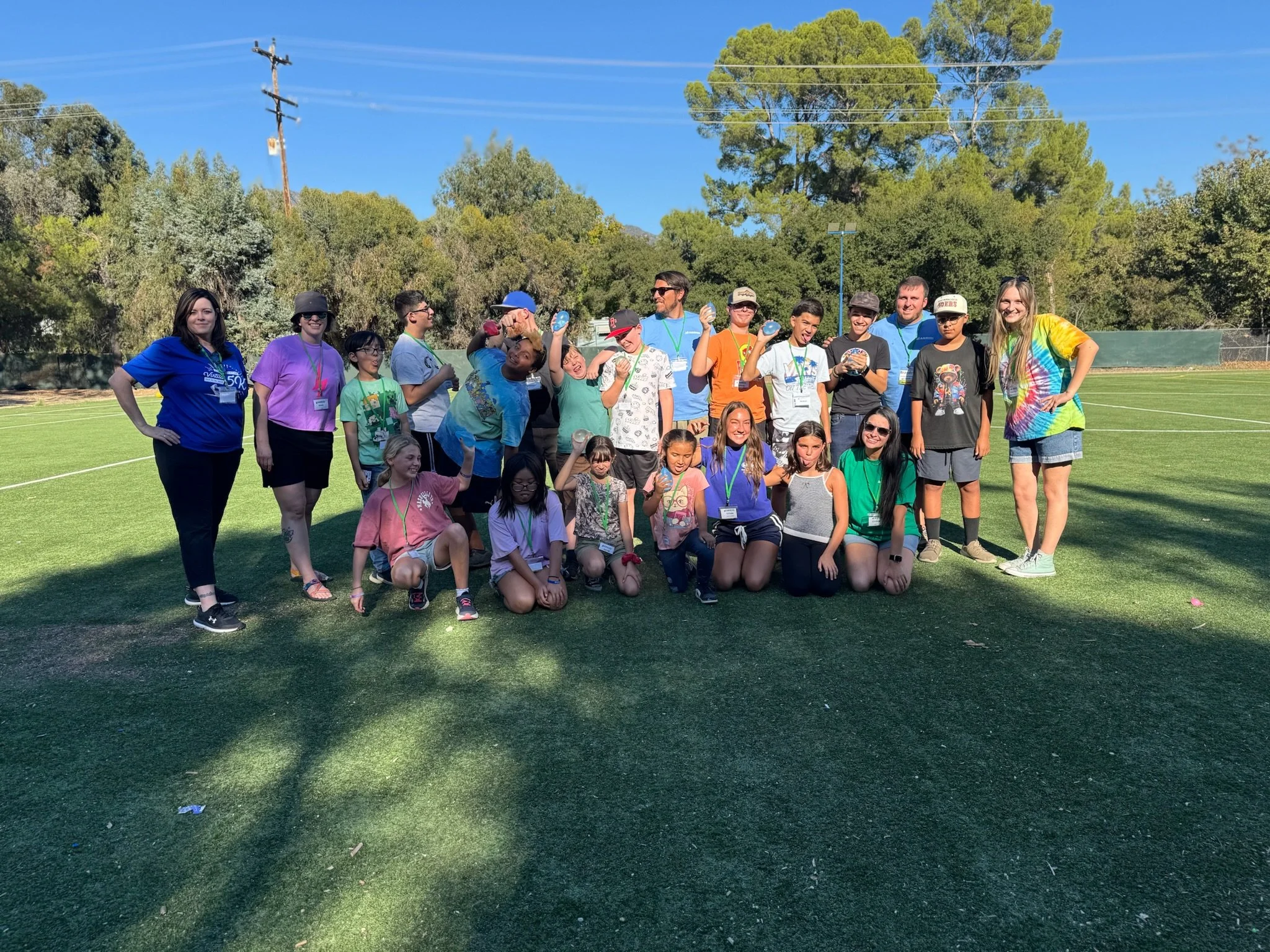 Group of children and adults standing on a grassy field, some holding medals, with trees and a clear blue sky in the background.