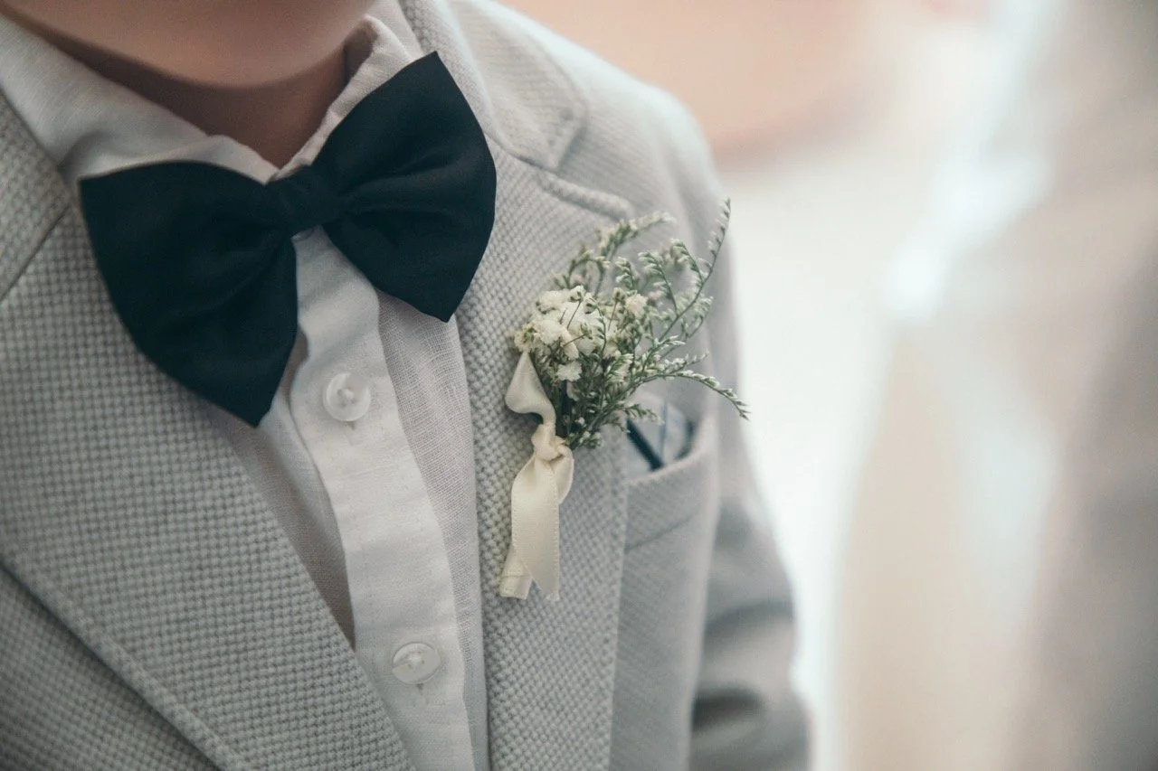 Close-up of a young man wearing a light grey checkered suit with a white shirt and a black bow tie, with a small bouquet of white flowers attached to his suit's pocket.