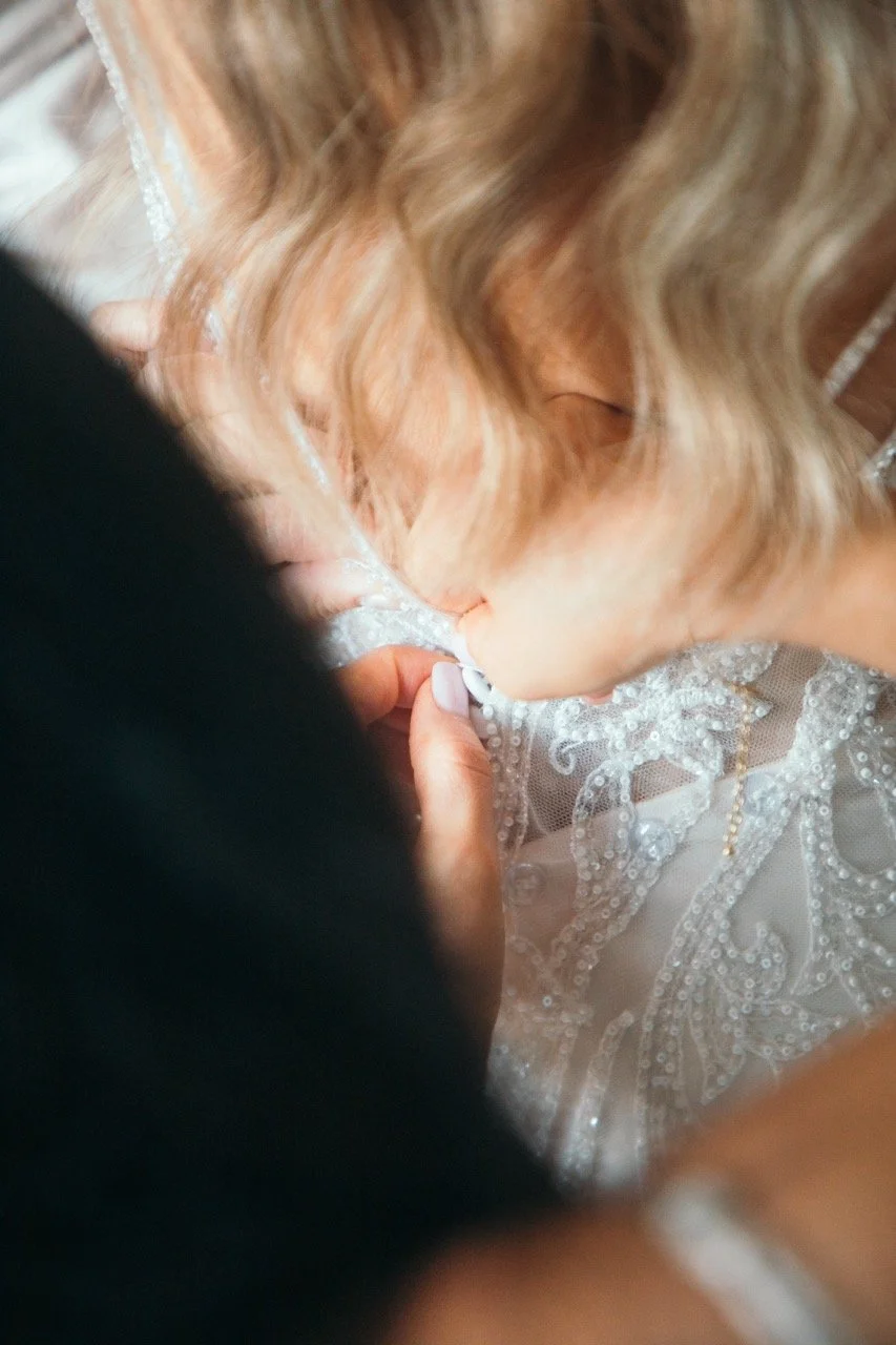 A person adjusting or fastening a lace or button on a white, ornate, beaded bride's wedding dress with blonde hair, seen from above.