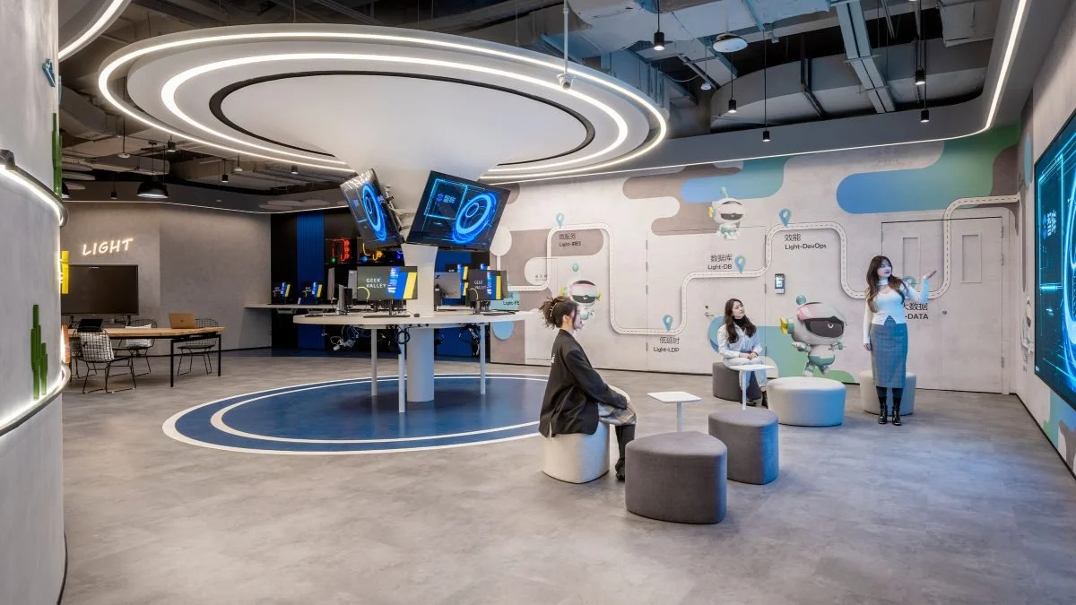 A modern technology showroom or office with a large digital screen wall, seating area with three women, and two monitors on a circular table, illuminated by circular ceiling lights.