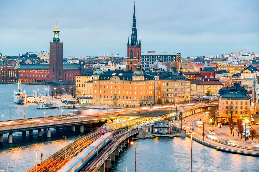 A cityscape of Stockholm, Sweden, with a river, historic buildings, a church with a tall spire, and a bridge with passing trains in the foreground. Learning Expedition