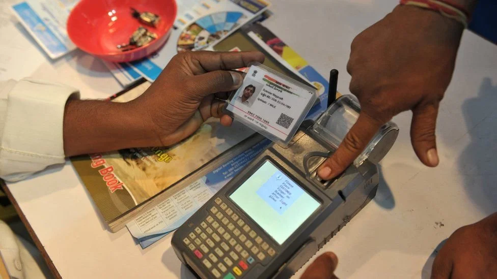 A person is holding a card near a point-of-sale machine while another person is pressing a button on the machine. The table has brochures, a red bowl, and some cards.