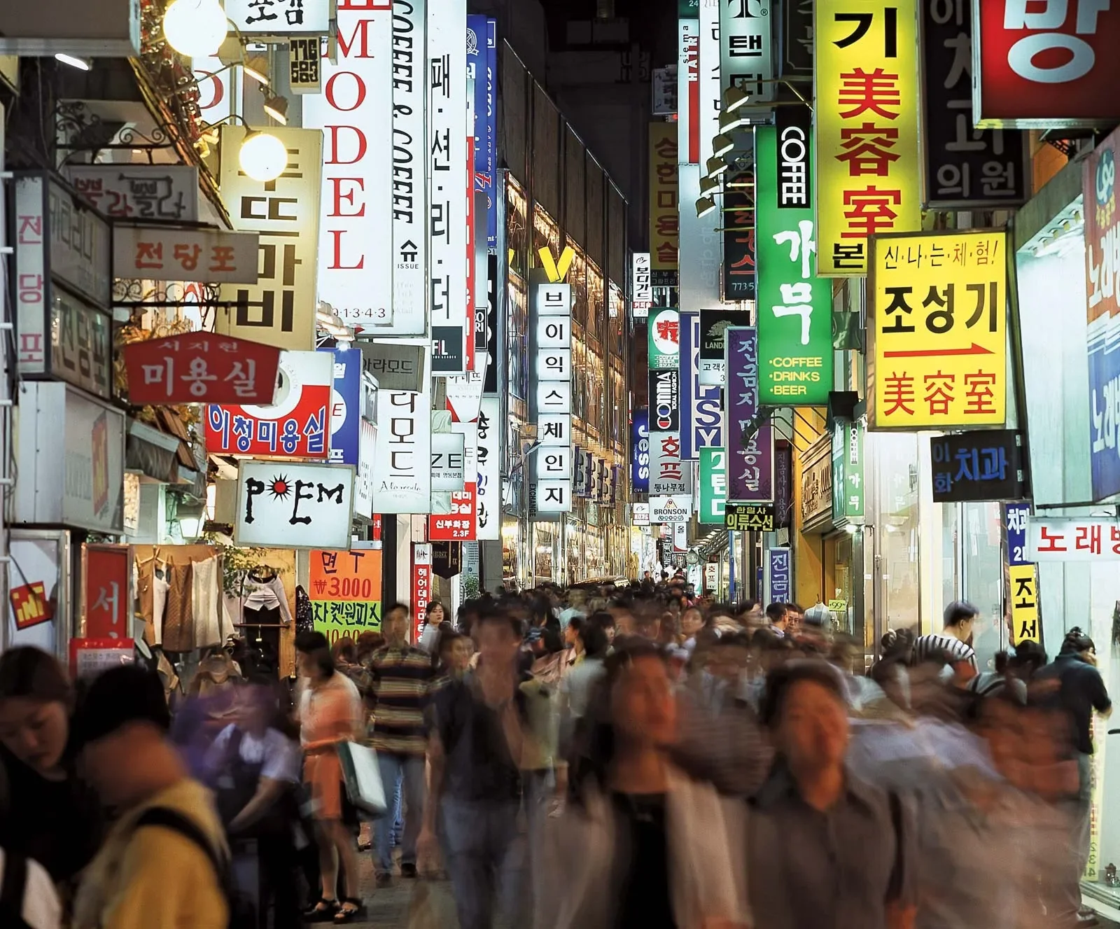 A busy city street at night with many colorful neon signs in Korean advertising various shops, restaurants, and services, densely packed with pedestrians walking and shopping. Learning Expedition