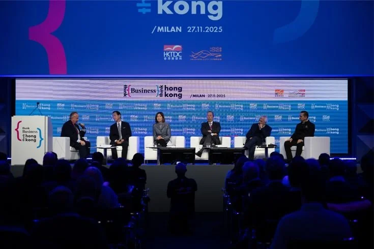 Panel discussion with six people on stage at a conference in Milan, held on November 27, 2025, with a large blue screen and banners displaying event details and logos in the background.