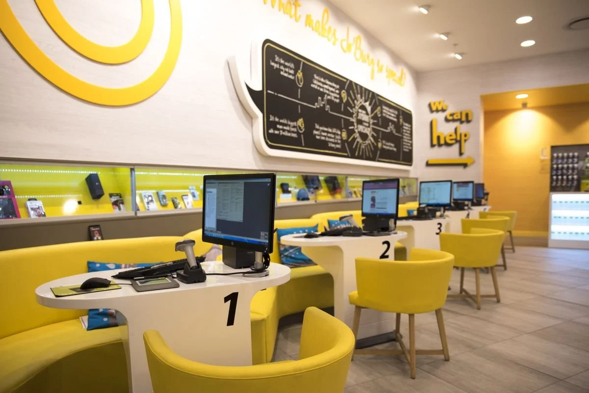 A woman wearing a yellow shirt and a face mask, holding a smartphone, standing in front of a convenience store with a 'OPEN 24 HOURS' sign and a menu in Chinese. She appears to be scanning or reading something on her phone.