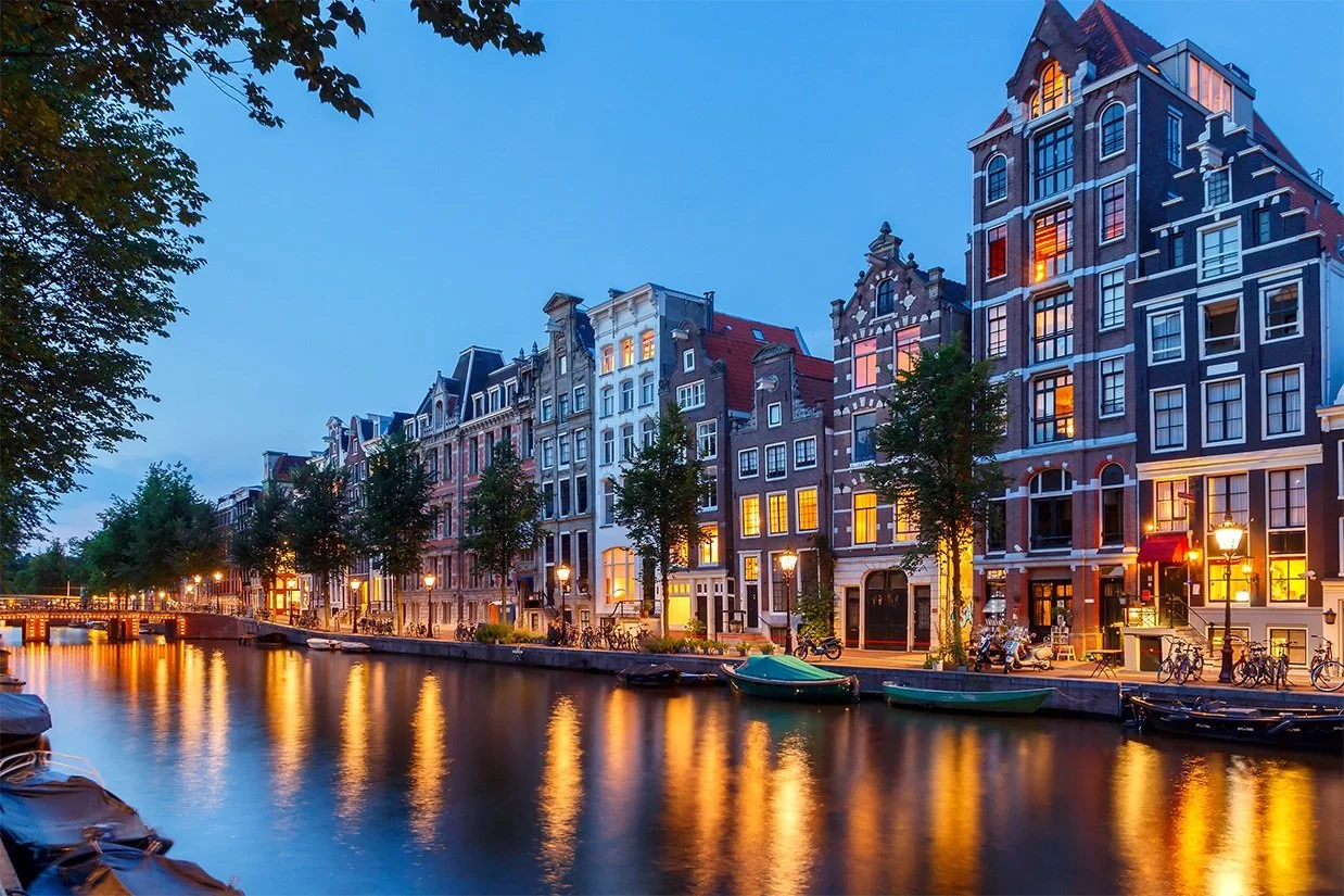 A canal in Amsterdam at dusk with colorful traditional Dutch buildings and boats along the water, illuminated by street lamps. Learning Expedition