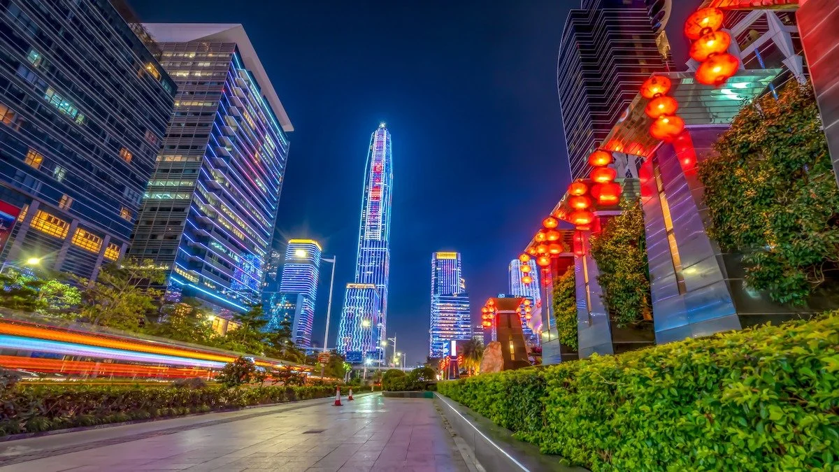 Night view of a modern city with tall, illuminated skyscrapers and red lanterns hanging on a building to the right. There are light trails from moving vehicles on the street in the foreground. Learning Expedition