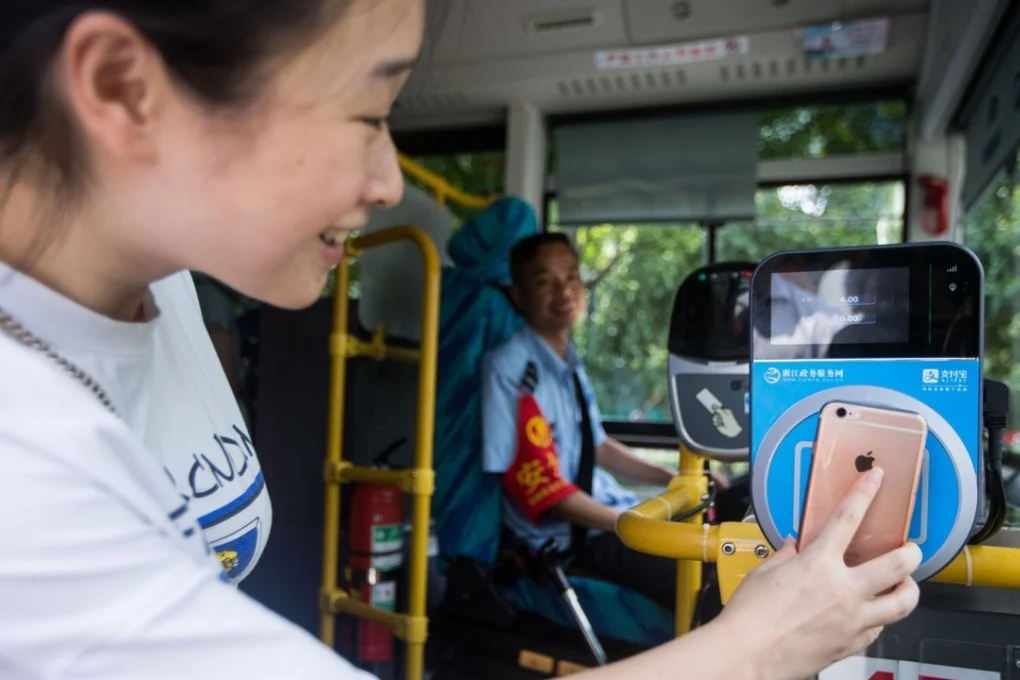 A woman using a contactless payment card on a bus, with a smiling bus driver in the background.