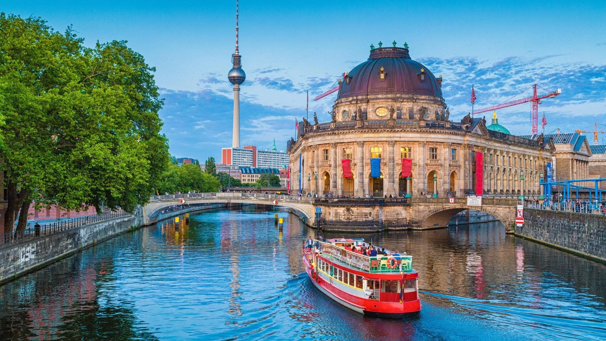 A scenic view of the Berlin City Palace with a boat cruising on the Spree River in front, lush green trees on the side, and the Fernsehturm tower in the background during sunset. Learning Expedition