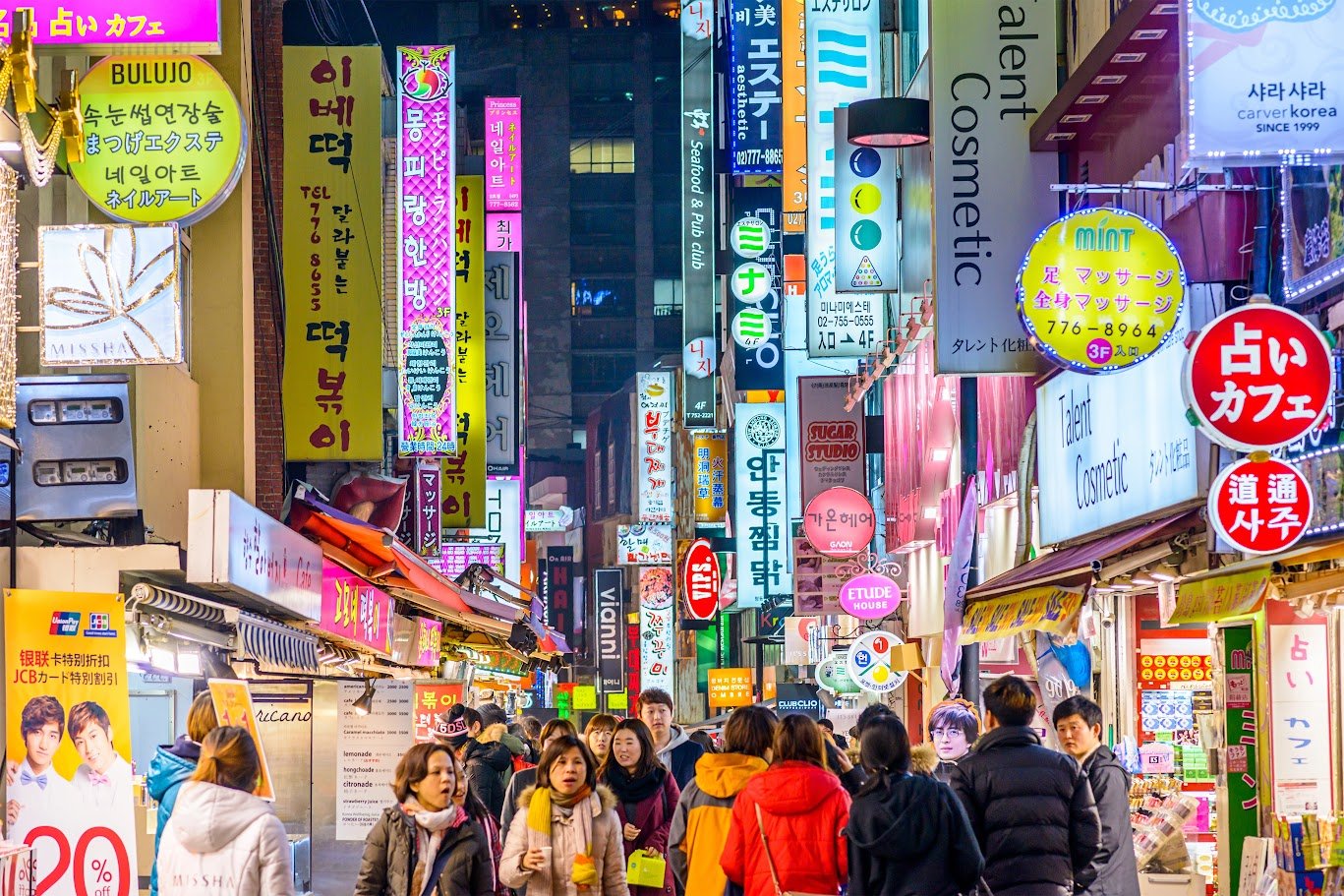 Shoppers on a neon-lit Seoul shopping street lined with Korean beauty and food stores, reflecting Hallyu demand