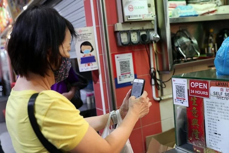 A woman wearing a yellow shirt and a face mask, holding a smartphone, standing in front of a convenience store with a 'OPEN 24 HOURS' sign and a menu in Chinese. She appears to be scanning or reading something on her phone.