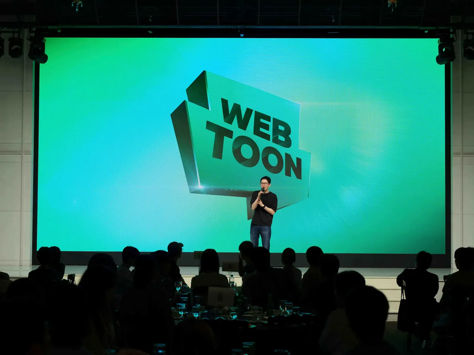 Speaker on stage before a large WEBTOON logo screen at a company event, illustrating the webtoon industry's growth