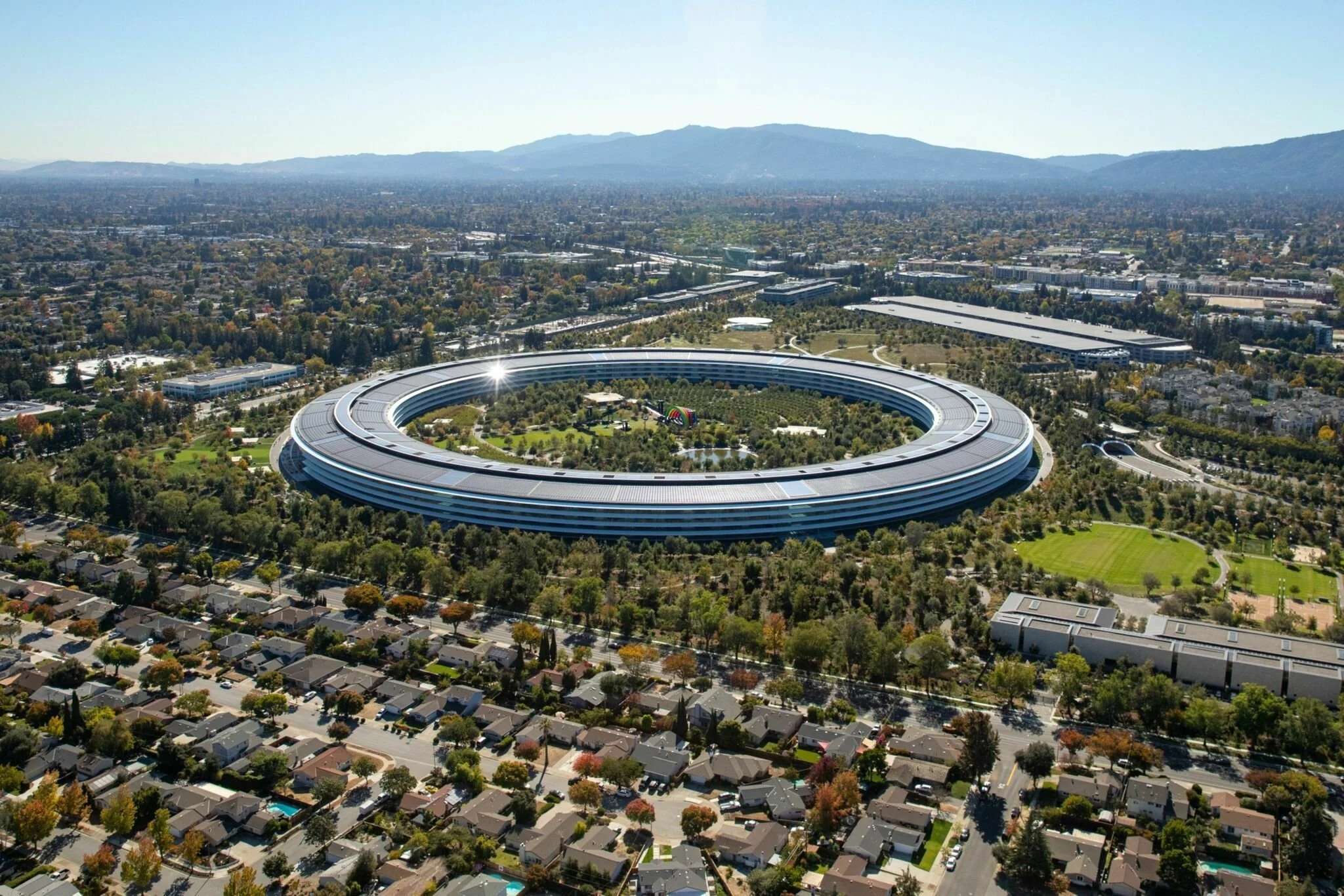 Aerial view of Apple Park, the circular corporate headquarters of Apple Inc., surrounded by trees, residential neighborhoods, and cityscape with mountains in the background. Learning Expedition