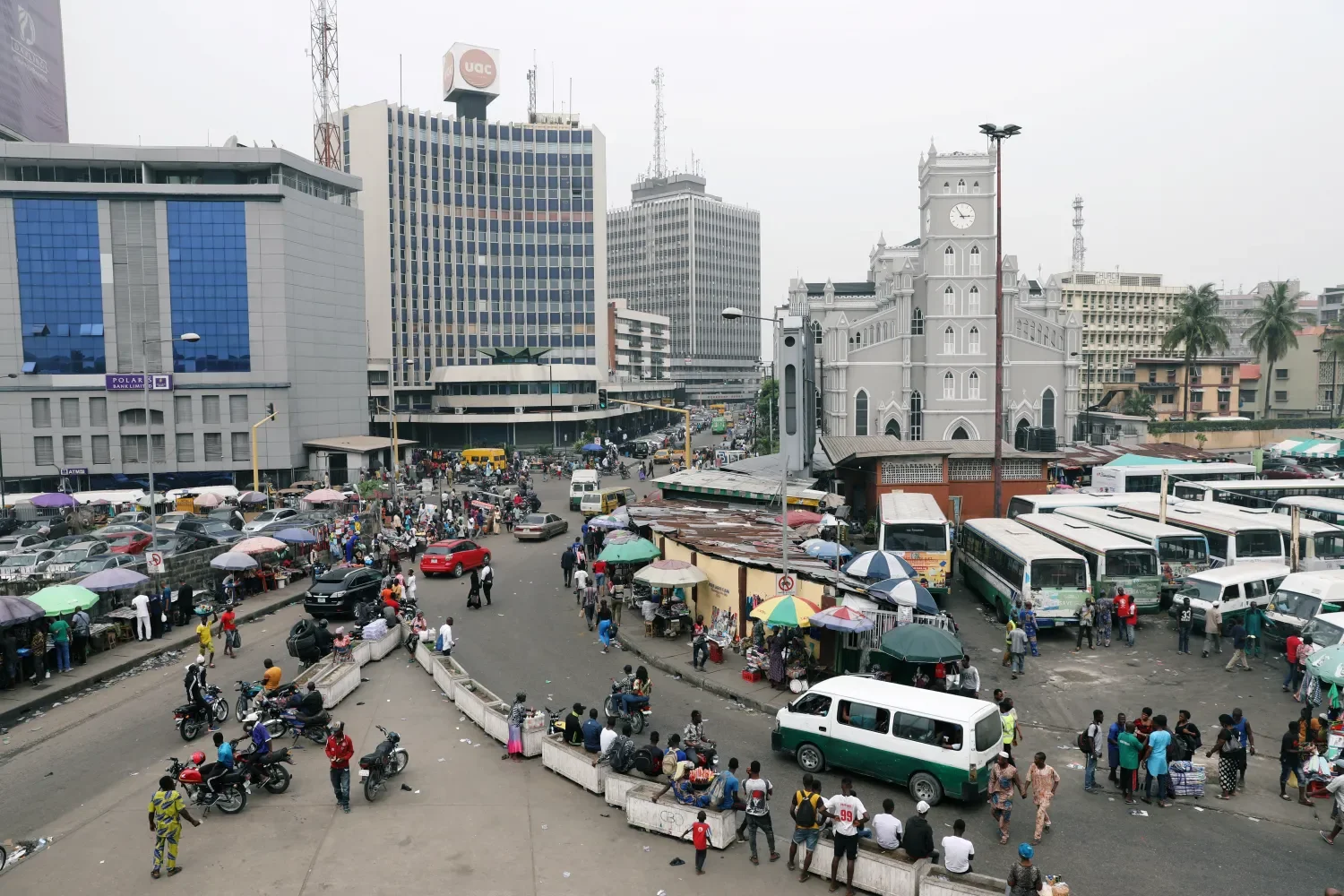 A busy city street scene with motorcycles, cars, buses, and pedestrians, surrounded by commercial buildings and an old white church with a clock tower. Learning Expedition