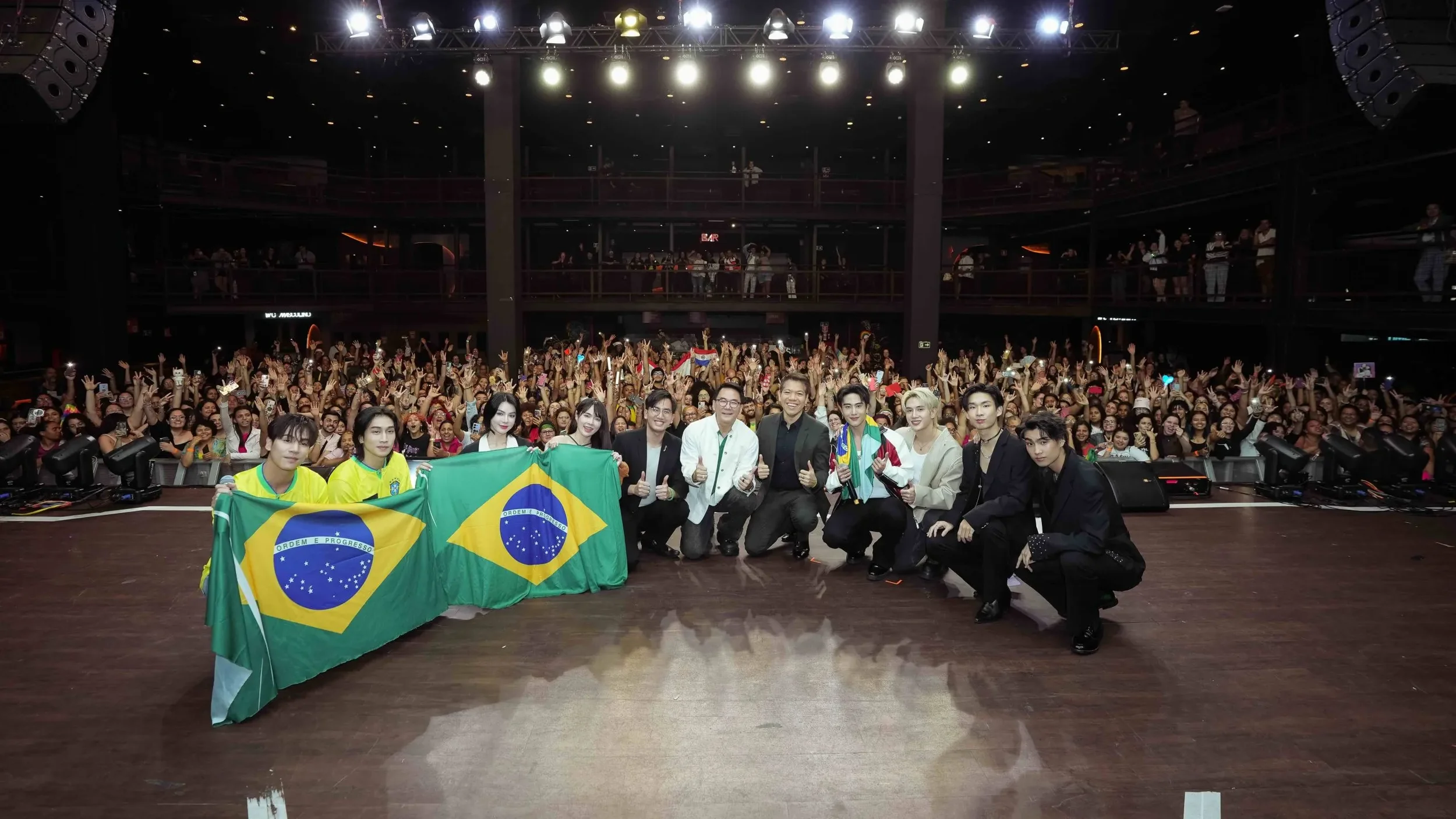 Thai BL actors and officials posing on stage with Brazilian flags before a cheering crowd at the Olá BL&GL Thailand in Brazil event