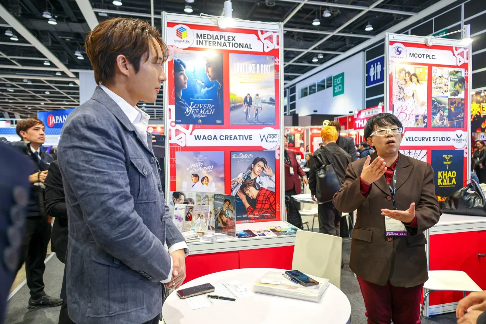 Exhibitor talking with a visitor at the Thailand Pavilion at FILMART 2025 in Hong Kong, beside Thai BL series posters
