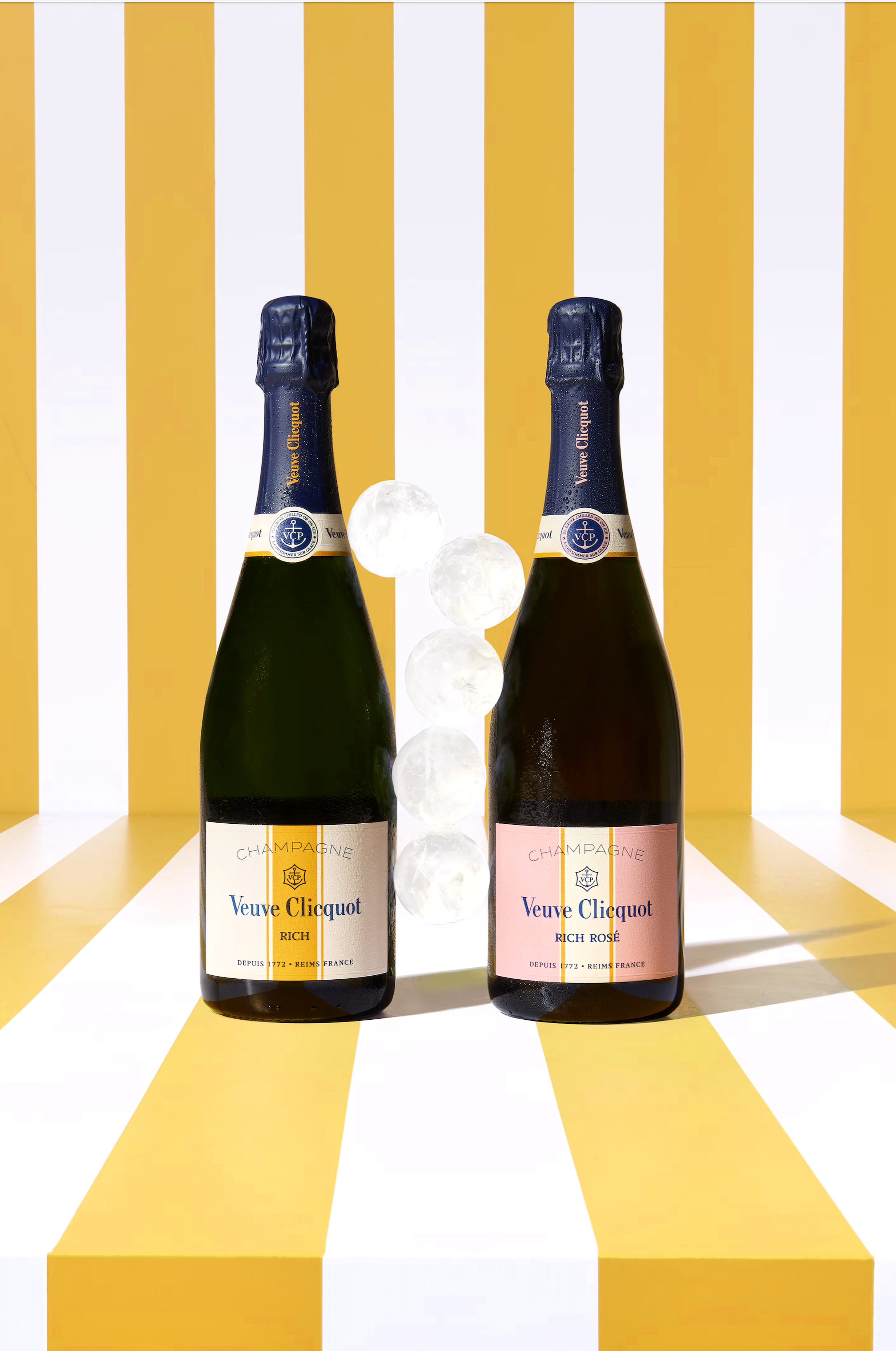 Two bottles of Veuve Clicquot champagne, one labeled 'Rich' and the other 'Rich Rosé,' standing on a yellow and white striped surface with a matching vertical striped background. There are three ice cubes placed between the bottles.