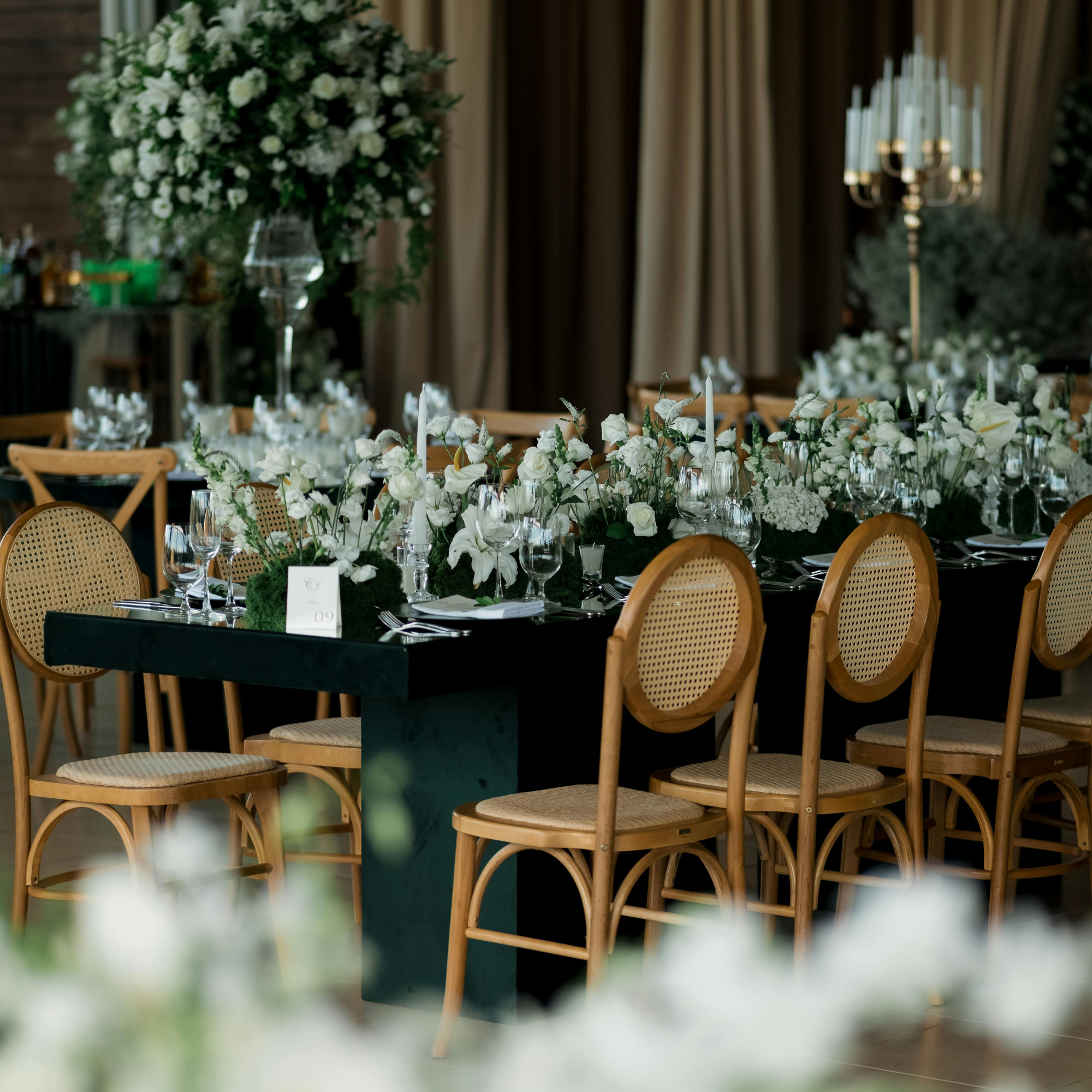 A blurred view of a decorated event space featuring a large floral arrangement with white flowers, a seating area with wooden chairs, and elegant chandeliers, likely prepared for a wedding or formal gathering.