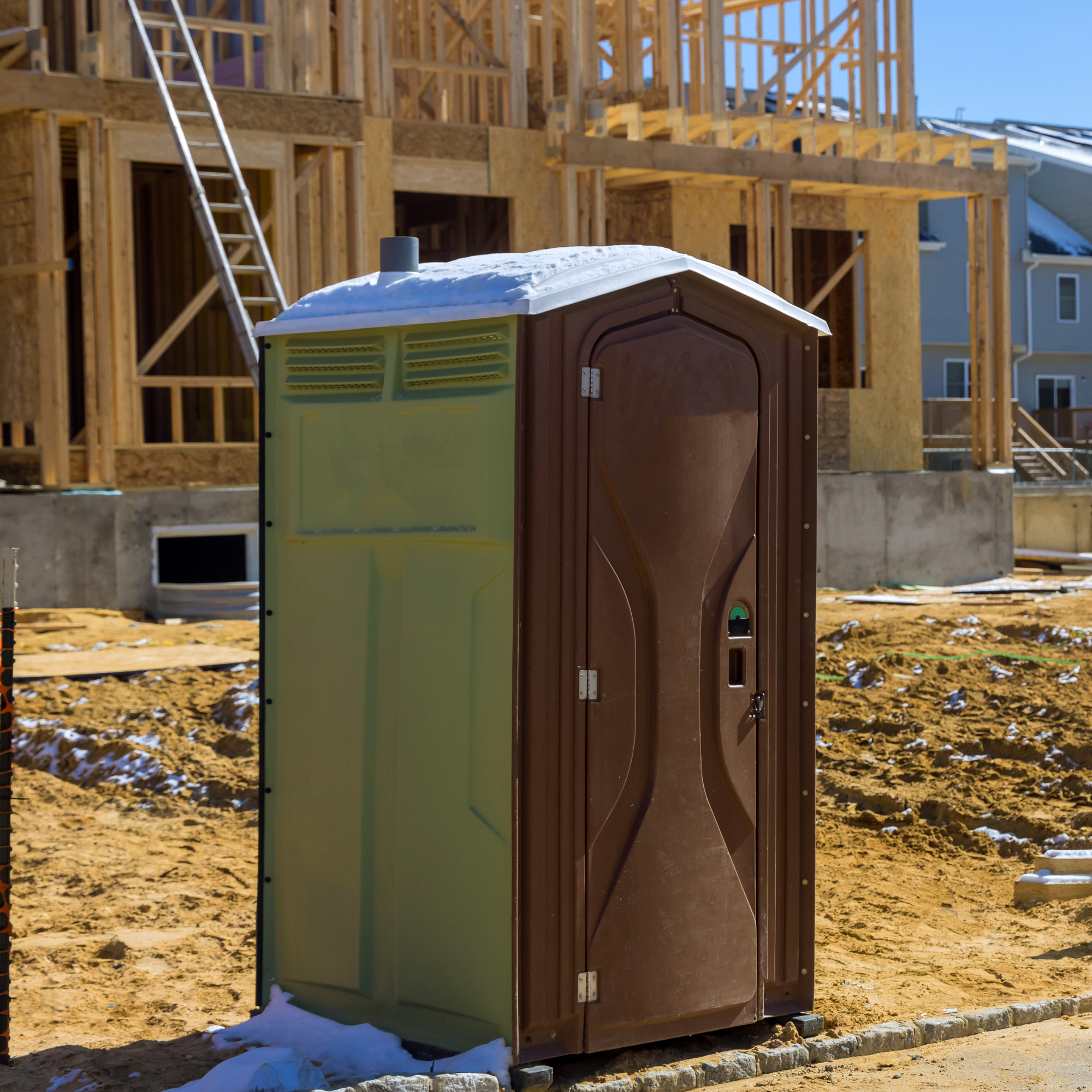 Construction site with a partially built wooden house, a ladder leaning against it, a portable toilet with snow on top, and piles of dirt on the ground.