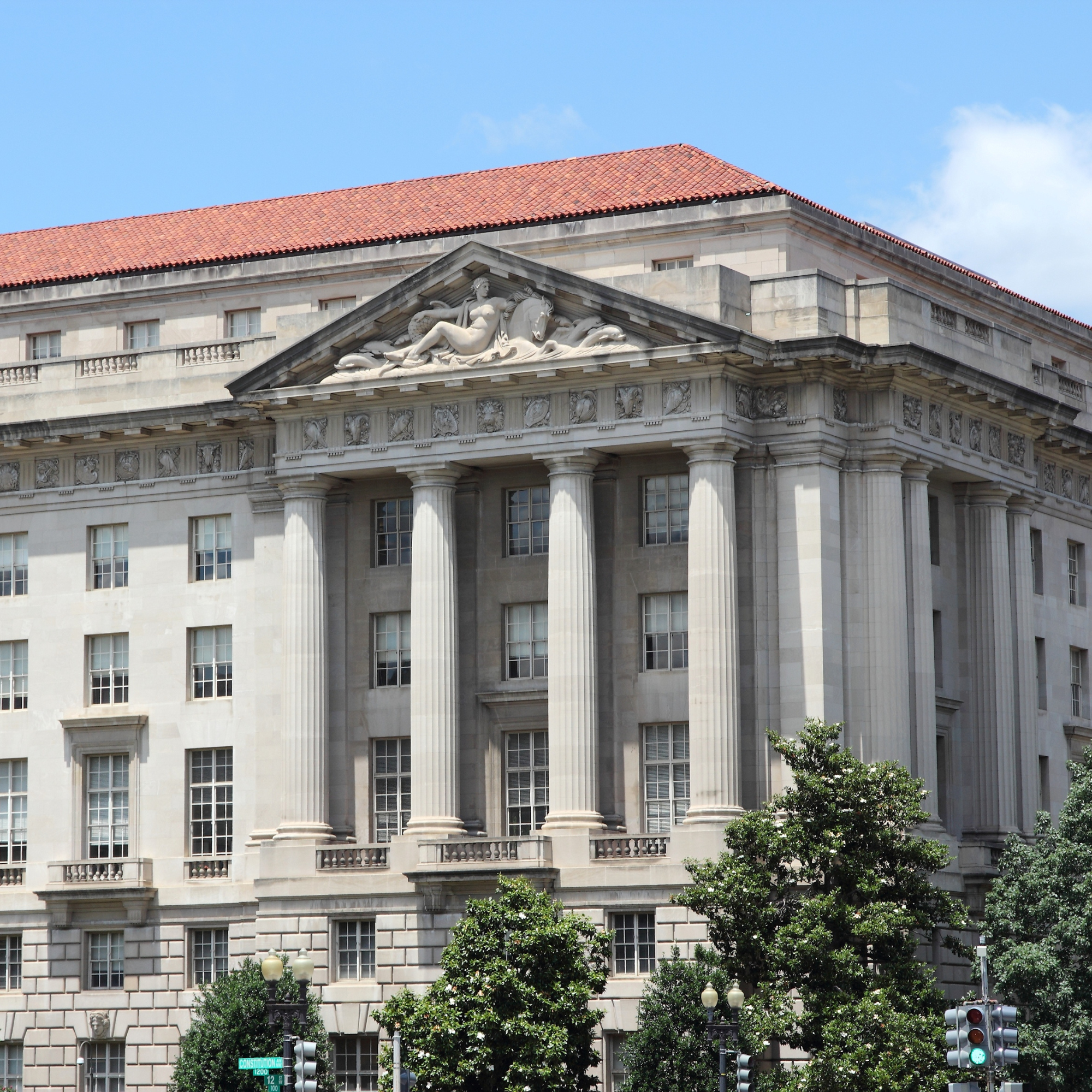 Photo of a classical building with a red tiled roof and an ornate sculpture of a reclining woman with a horse on the pediment. There are trees and traffic lights in the foreground.