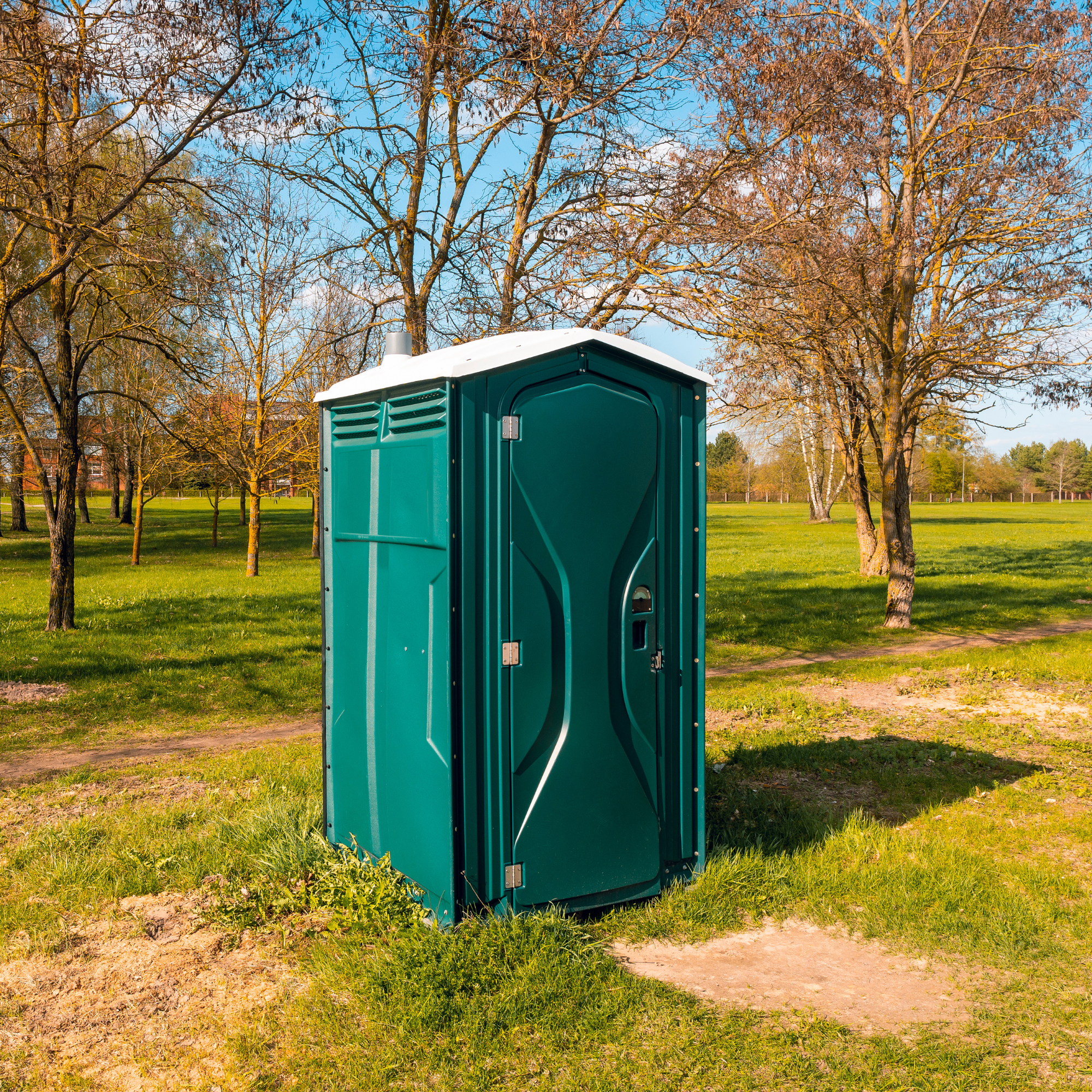A portable toilet situated in a park with green grass and trees with no leaves, under a clear blue sky.