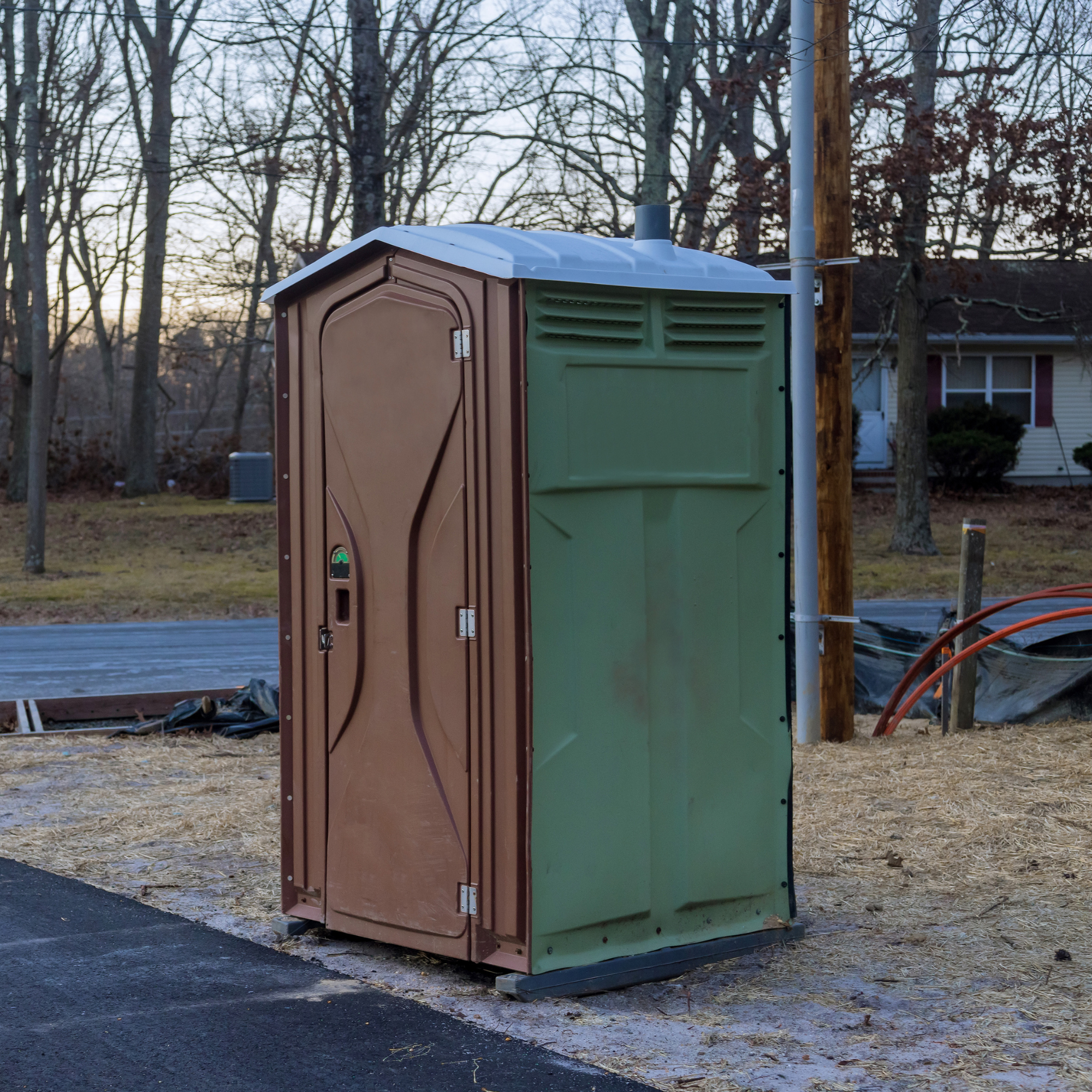 A portable toilet in a natural outdoor setting with leafless trees and a house visible in the background.