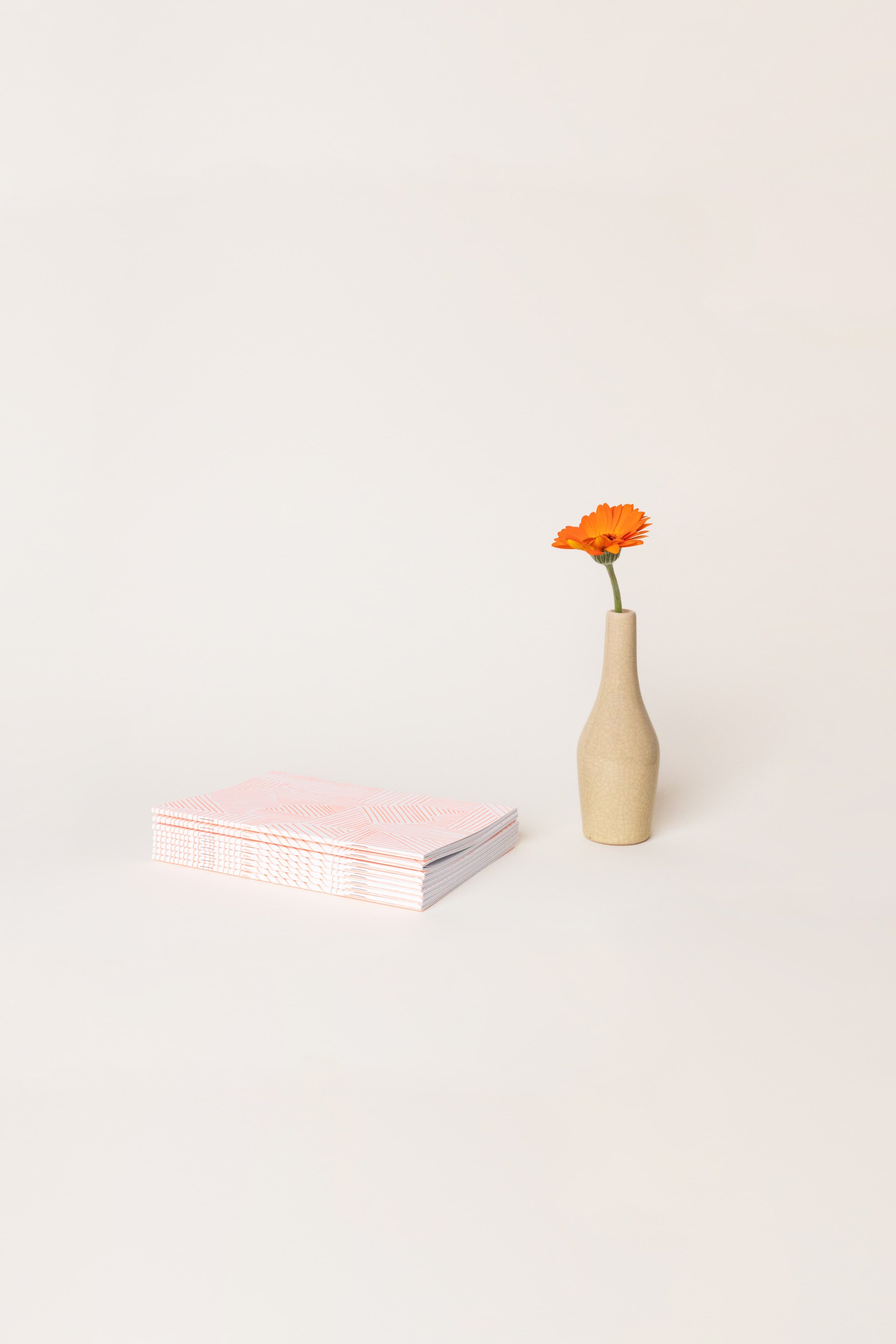 Une seule fleur orange dans un vase beige se trouve à côté d’une pile de papiers roses sur une surface blanche minimale.